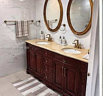 Dual-sink vanity with a stone countertop, ornate dark wood cabinetry, brass-toned fixtures, and oval wall-mounted mirrors set against large-format tiled walls