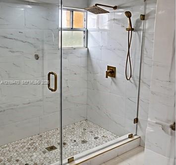 Walk-in shower enclosure featuring floor-to-ceiling white tile with charcoal veining, pebble-mosaic floor, glass door, and antique brass fixtures including a rain showerhead and handheld wand