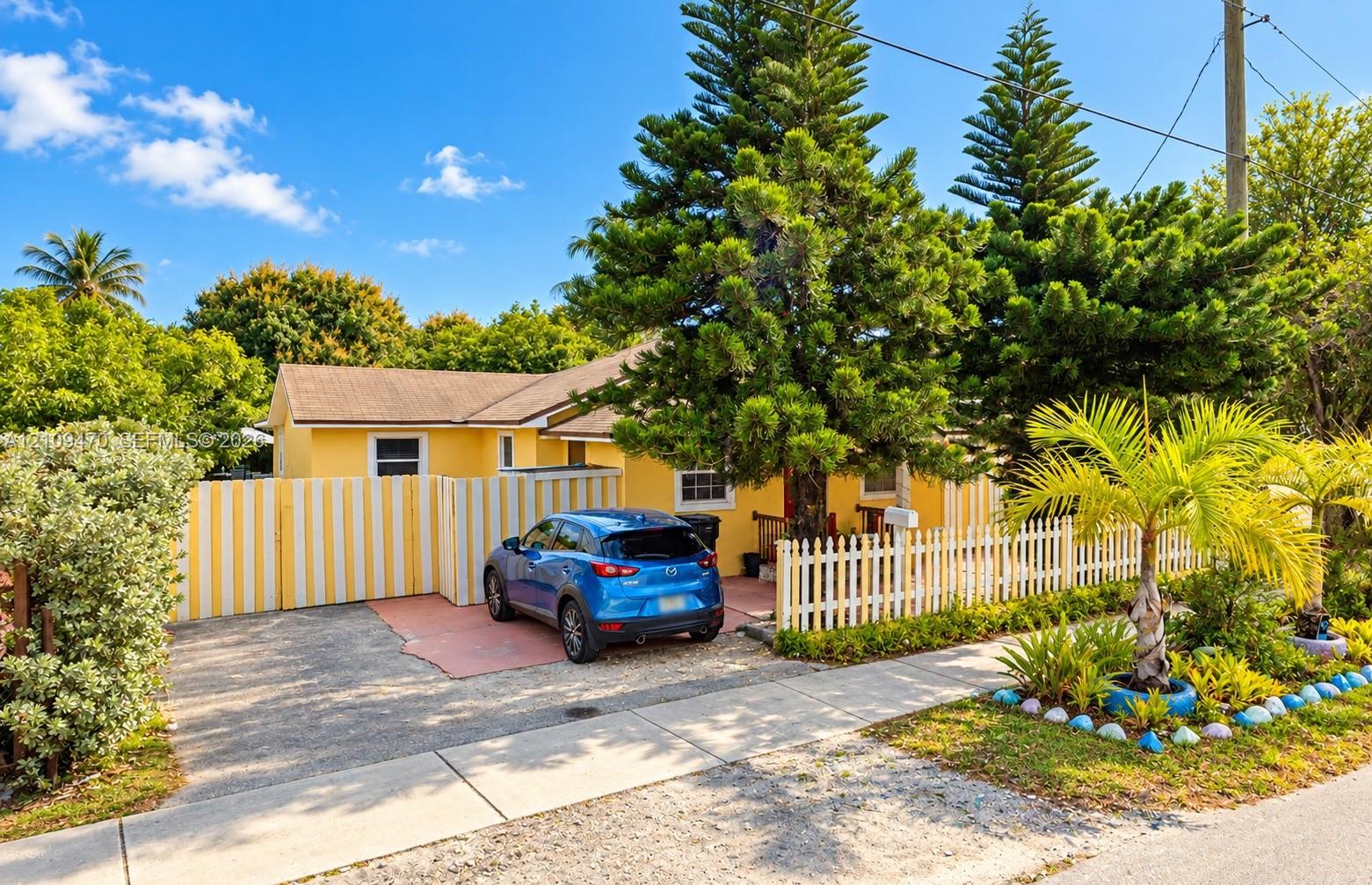 Single-story exterior featuring a vibrant yellow facade, paved driveway, picket fencing, mature pine trees, and established tropical landscaping Single-story exterior featuring a vibrant yellow facade, paved driveway, picket fencing, mature pine trees, and established tropical landscaping