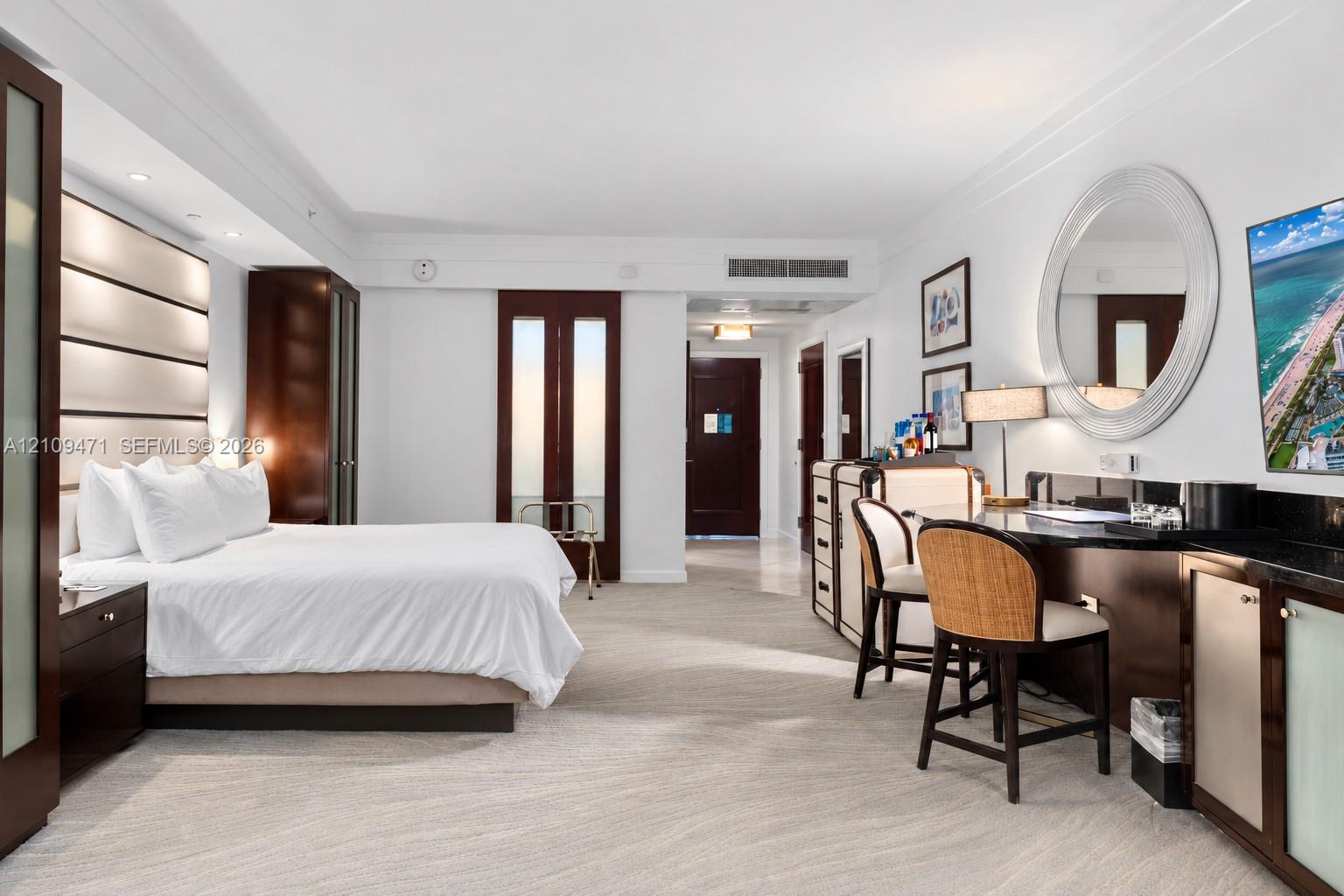 Spacious room featuring a wall-mounted panel headboard, dark wood-finish cabinetry, a built-in stone countertop desk, a circular wall mirror, and neutral-toned carpeting Spacious room featuring a wall-mounted panel headboard, dark wood-finish cabinetry, a built-in stone countertop desk, a circular wall mirror, and neutral-toned carpeting