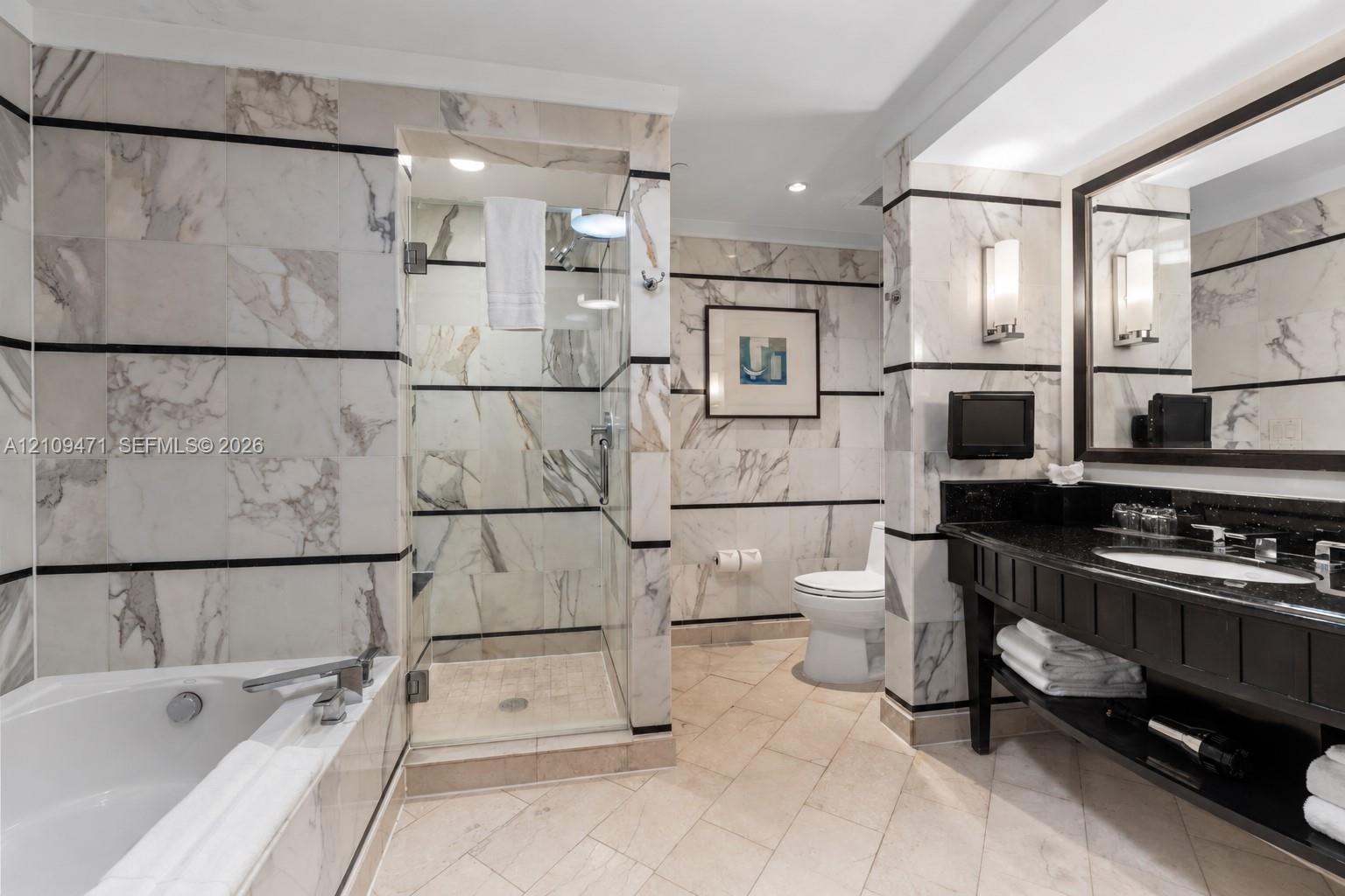 Bathroom featuring full-height natural stone wall tiling with horizontal accent banding, a glass-enclosed shower, a deep soaking tub, and a vanity with a dark stone countertop and integrated sink Bathroom featuring full-height natural stone wall tiling with horizontal accent banding, a glass-enclosed shower, a deep soaking tub, and a vanity with a dark stone countertop and integrated sink