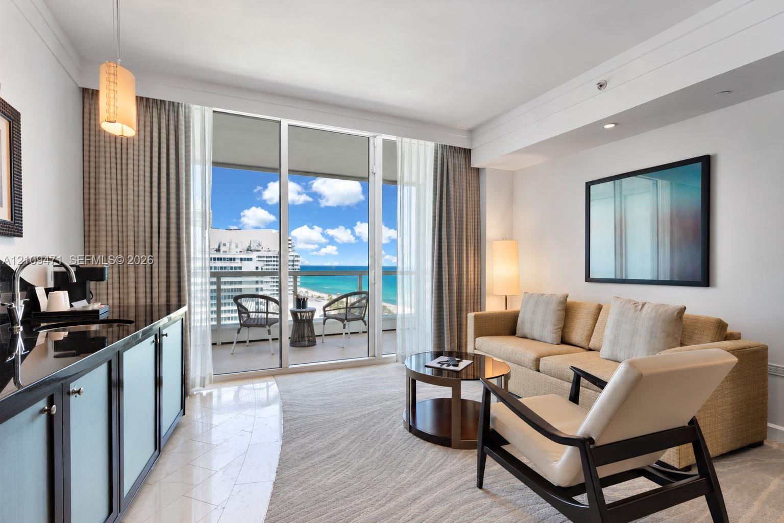 Living area featuring floor-to-ceiling glass sliding doors, private balcony with ocean frontage, wet bar with dark stone countertop, and polished tile flooring Living area featuring floor-to-ceiling glass sliding doors, private balcony with ocean frontage, wet bar with dark stone countertop, and polished tile flooring