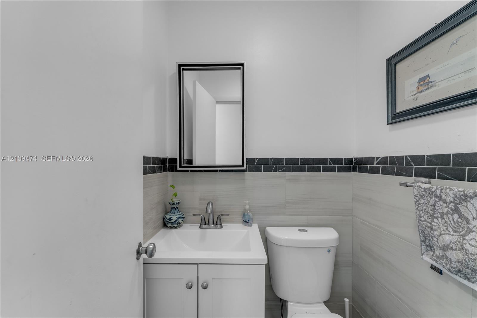 White vanity with a rectangular sink, brushed nickel faucet, wall-mounted mirror with a black frame, and gray stone-finish wall tile featuring a black linear tile border