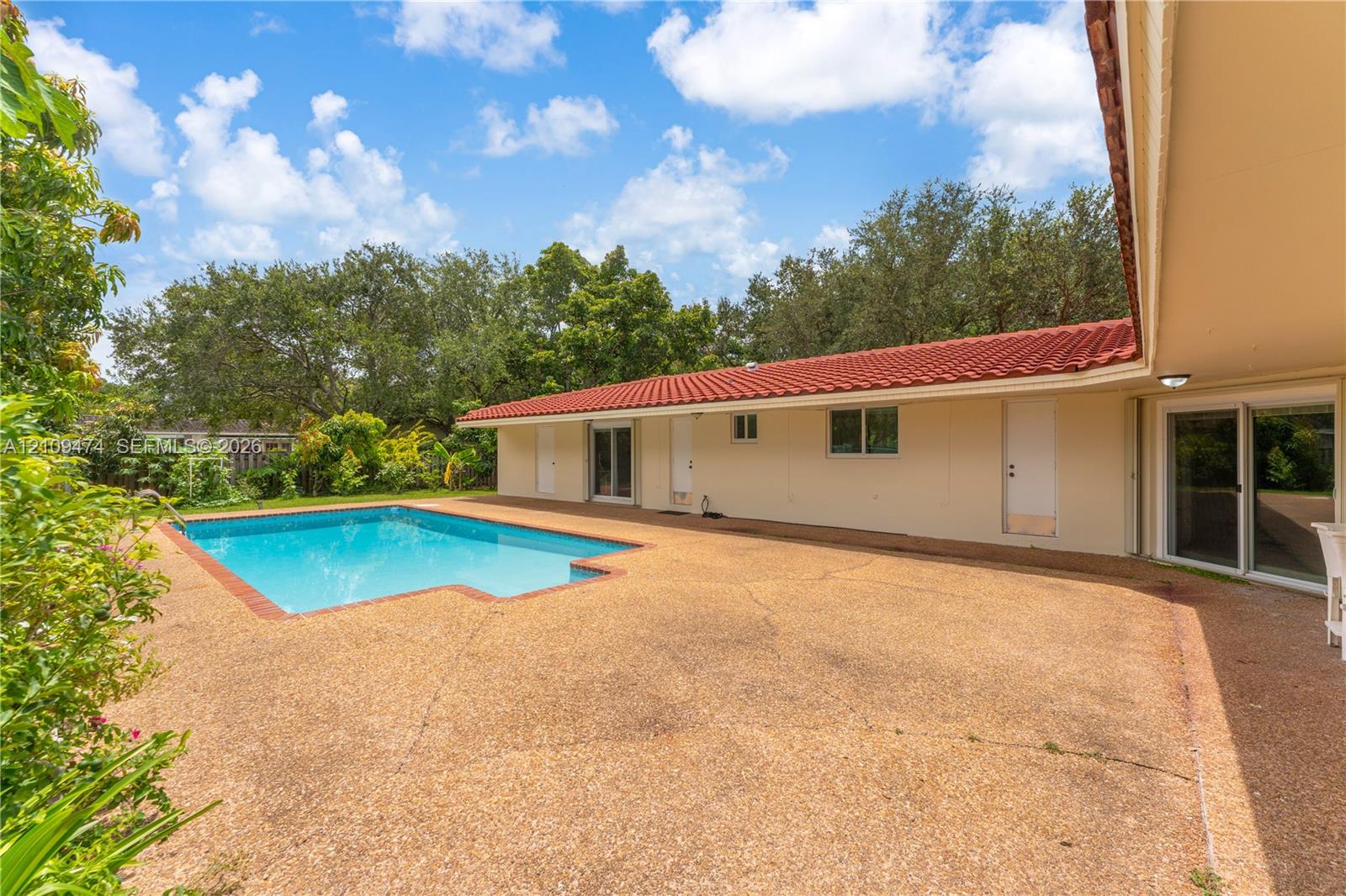 In-ground swimming pool featuring a red brick border, expansive aggregate patio, red tile roof, and multiple glass sliding door entries