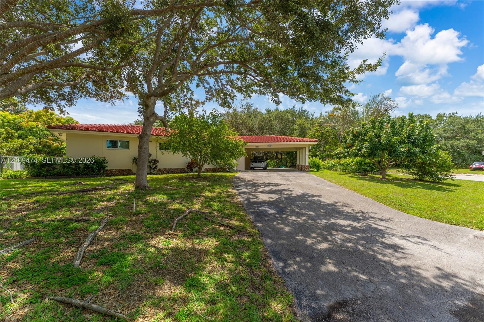 Single-story exterior featuring a red tile roof, covered carport, and asphalt driveway situated on a landscaped lot with mature trees