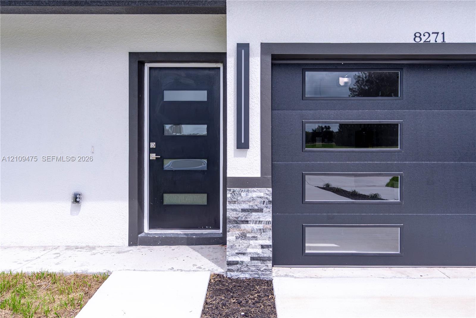 Modern exterior featuring a smooth-stucco facade, stacked stone accent column, dark-finish entry door with horizontal glass inserts, matching panel garage door with rectangular windows, and a vertical linear wall-mounted light fixture
