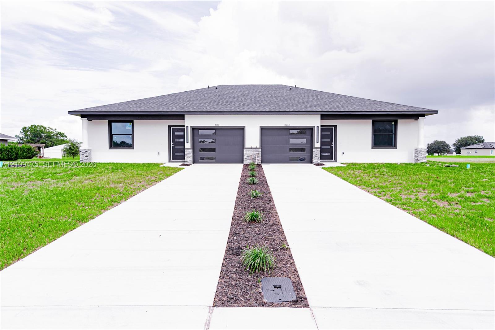 Contemporary duplex featuring a light-colored stucco exterior, dual attached garages with modern glass-paneled doors, stacked stone columns, and a concrete driveway with a central mulched garden bed