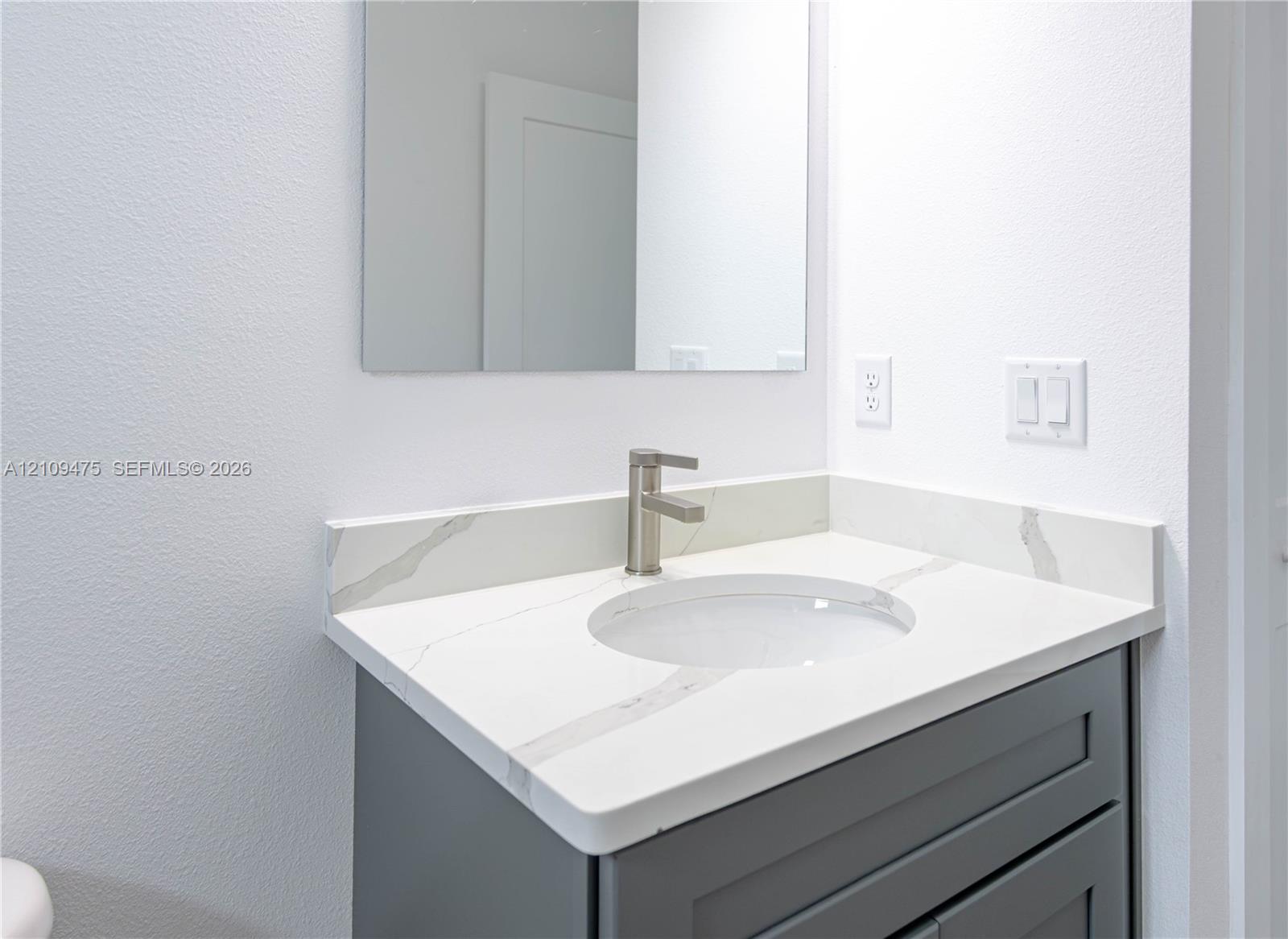 Vanity featuring a grey cabinet, white stone-composite countertop with veining, undermount sink, brushed nickel faucet, and frameless mirror