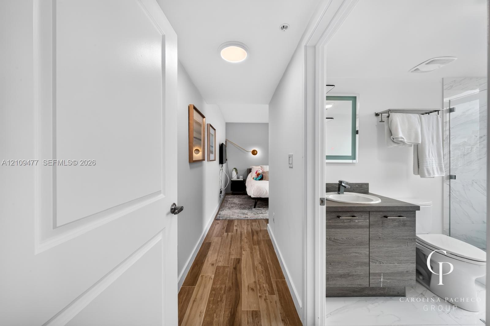 Wood-finish flooring throughout, bathroom featuring a vanity with dark-tone cabinetry, integrated sink, and an illuminated mirror, and a walk-in shower with a glass enclosure