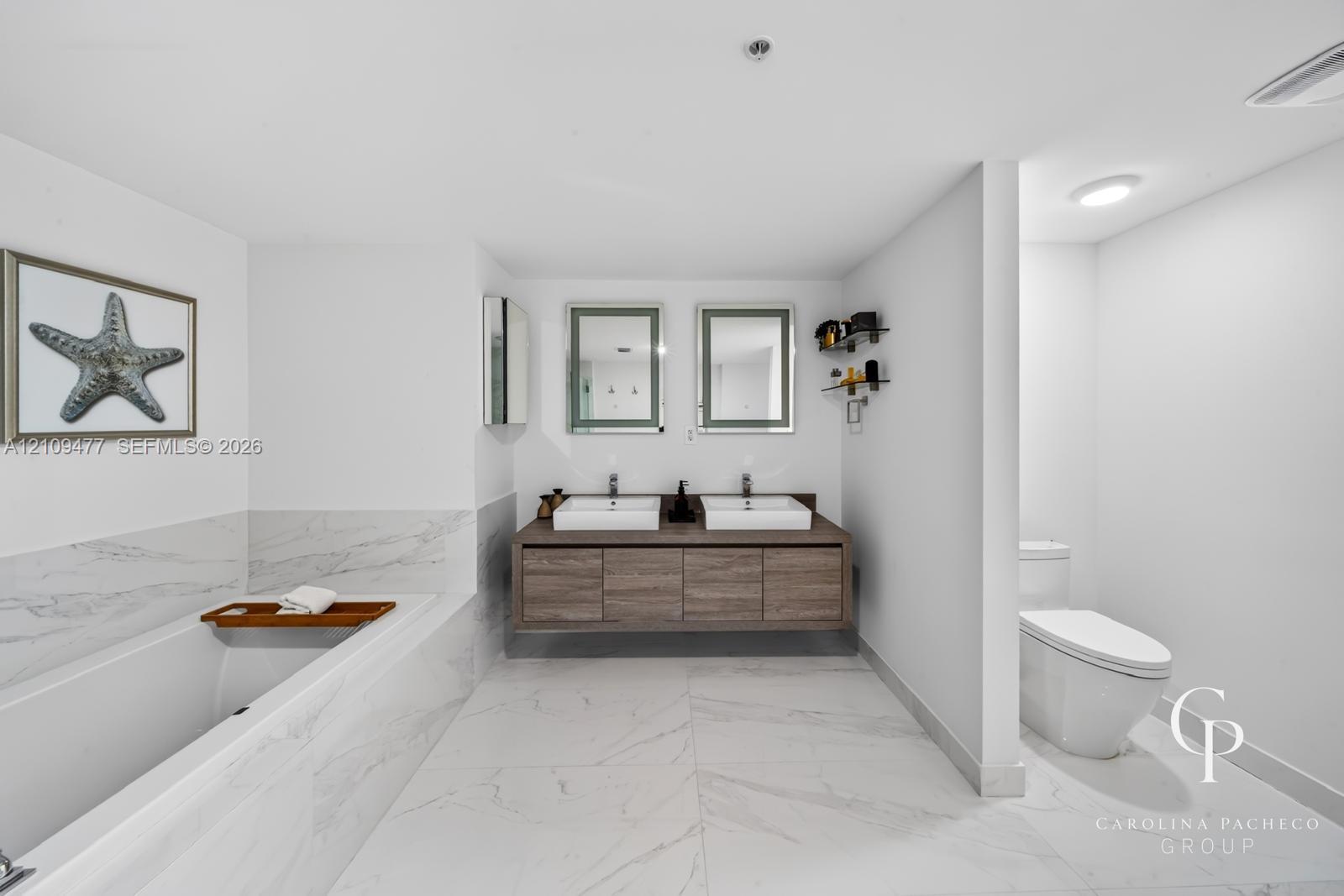 Floating dual-sink vanity with wood-finish cabinetry, rectangular vessel sinks, wall-mounted rectangular mirrors, soaking tub, and large-format tile flooring