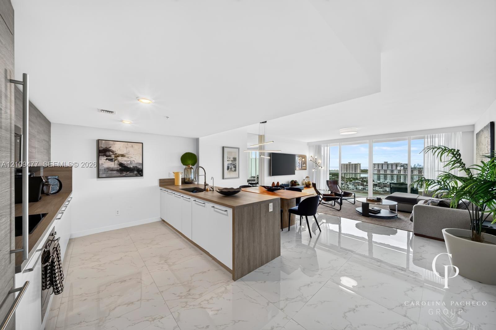 Open-concept kitchen featuring an island with wood-finish cabinetry, white flat-panel lower cabinets, stone-surfaced countertops, and polished tile flooring extending throughout the main living space, finished with floor-to-ceiling windows
