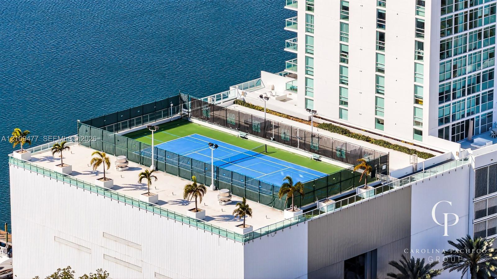 Rooftop tennis court featuring professional-grade blue surfacing, protective perimeter netting, integrated lighting, and decorative planters
