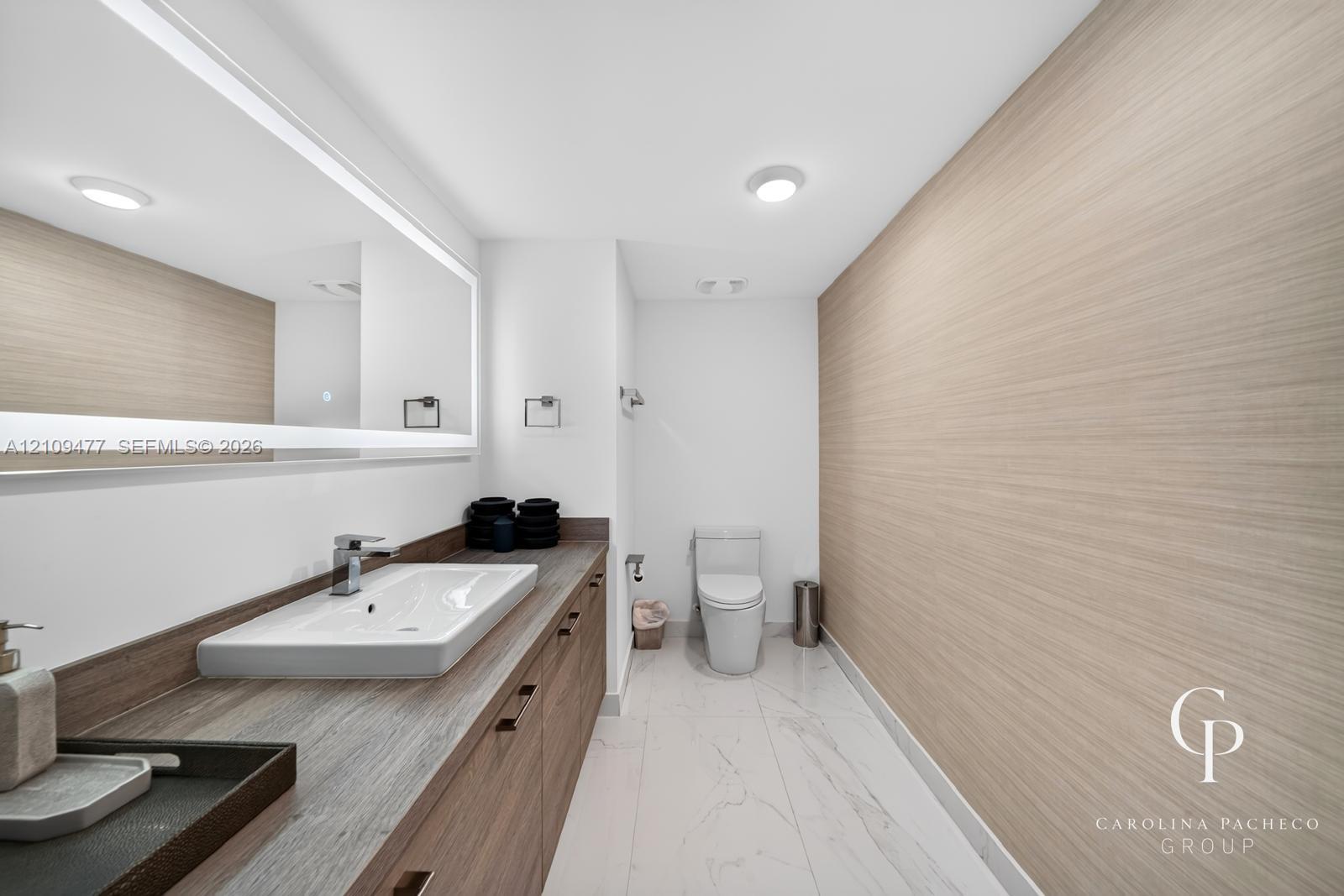Bathroom featuring a wood-finish vanity with a rectangular vessel sink, integrated lighting mirror, stone-finish countertop, and large-format tile flooring
