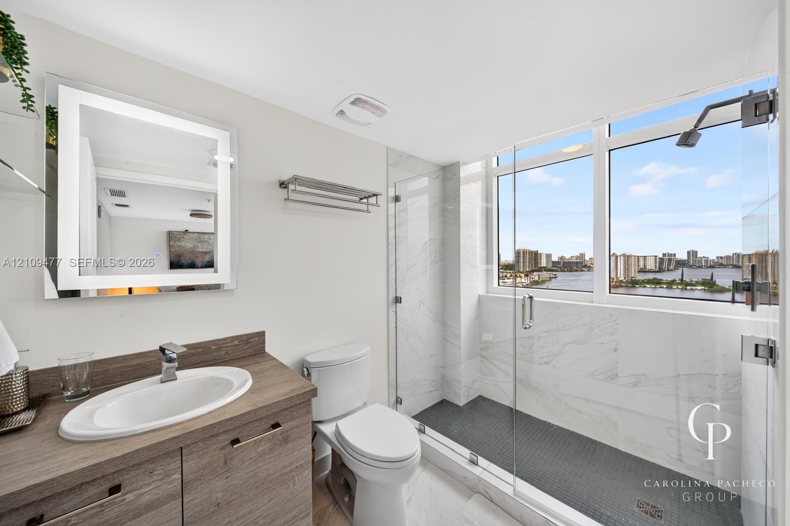 Bathroom featuring a wood-finish vanity with an integrated basin, a backlit mirror, and a walk-in shower with stone-tile surround, dark floor tiling, and glass enclosure