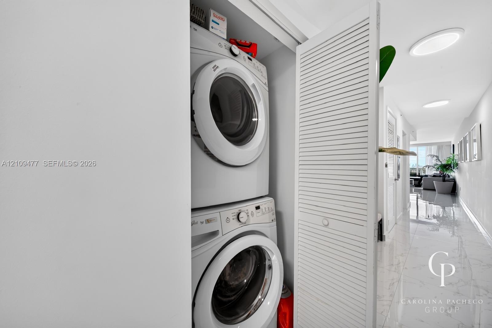 Laundry closet featuring stacked front-load appliances and louvered folding doors, accented by polished stone-finish flooring and recessed lighting