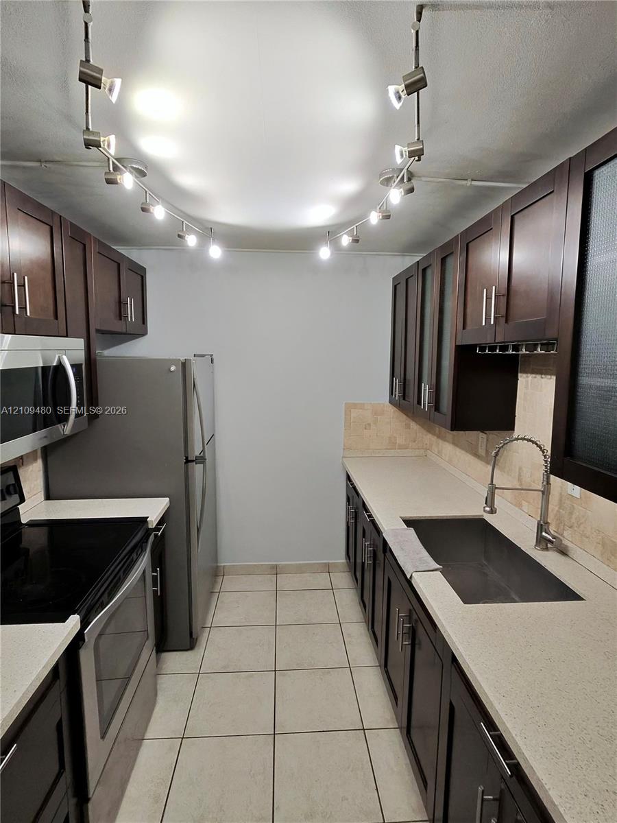 Kitchen featuring dark wood-finish cabinetry, solid surface countertops, stainless steel appliances, ceramic tile flooring, and adjustable track lighting