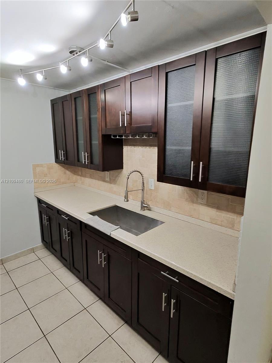 Dark-toned shaker cabinetry featuring frosted glass door inserts, stone-finish countertops, stainless steel undermount sink, tiled backsplash, and multi-light track lighting