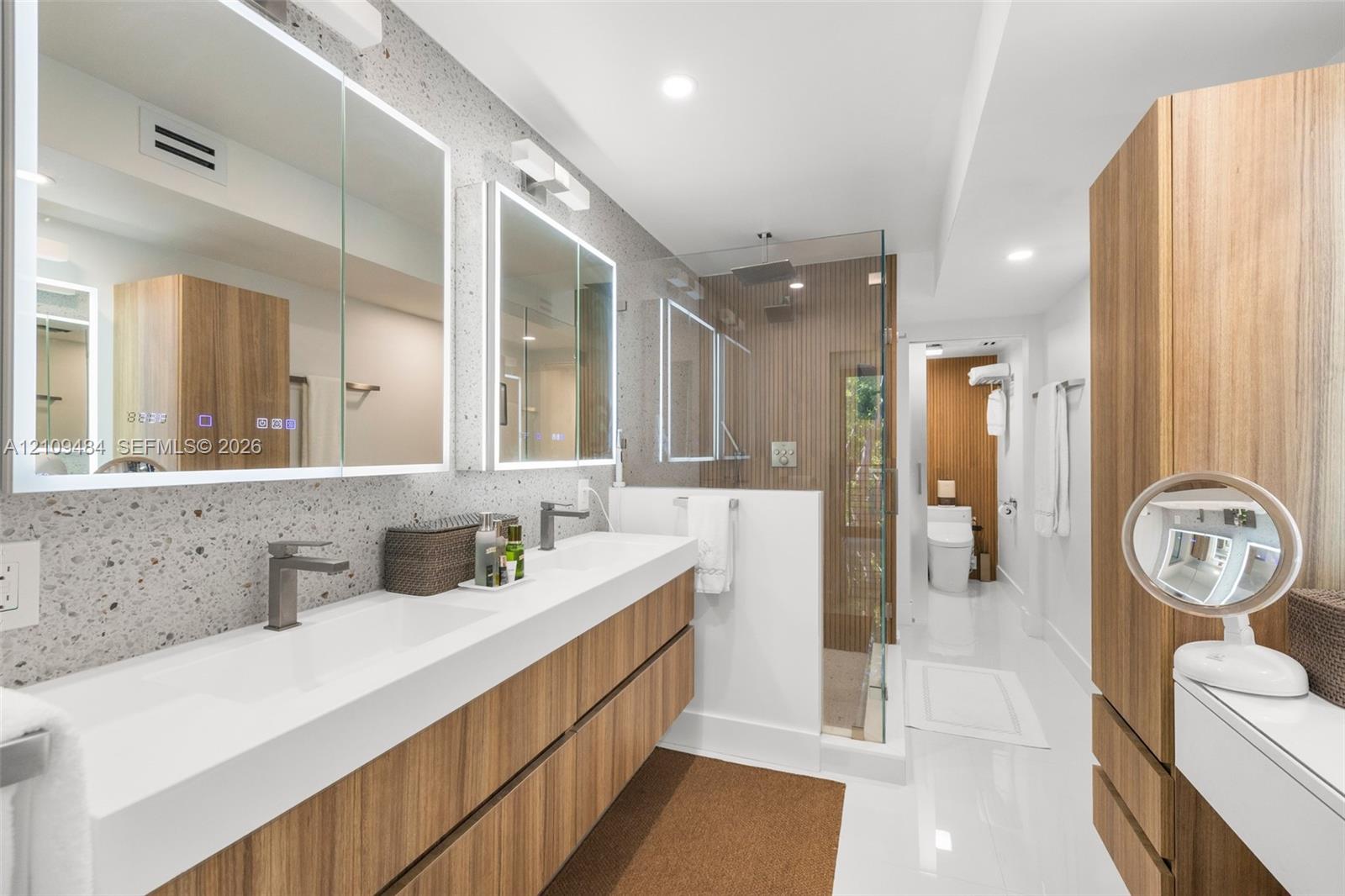 Floating dual-vanity with integrated sinks and wood-finish cabinetry, illuminated mirrored medicine cabinets, terrazzo-style wall surfacing, walk-in glass shower enclosure, and floor-to-ceiling wood-finish storage cabinetry