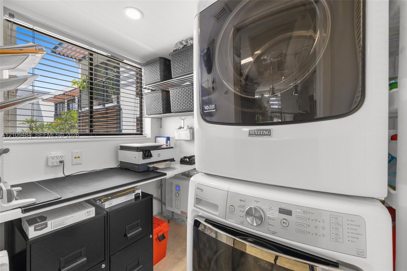 Laundry area featuring a stacked washer and dryer unit, built-in surface counter, metal wire shelving, and a window with horizontal blinds
