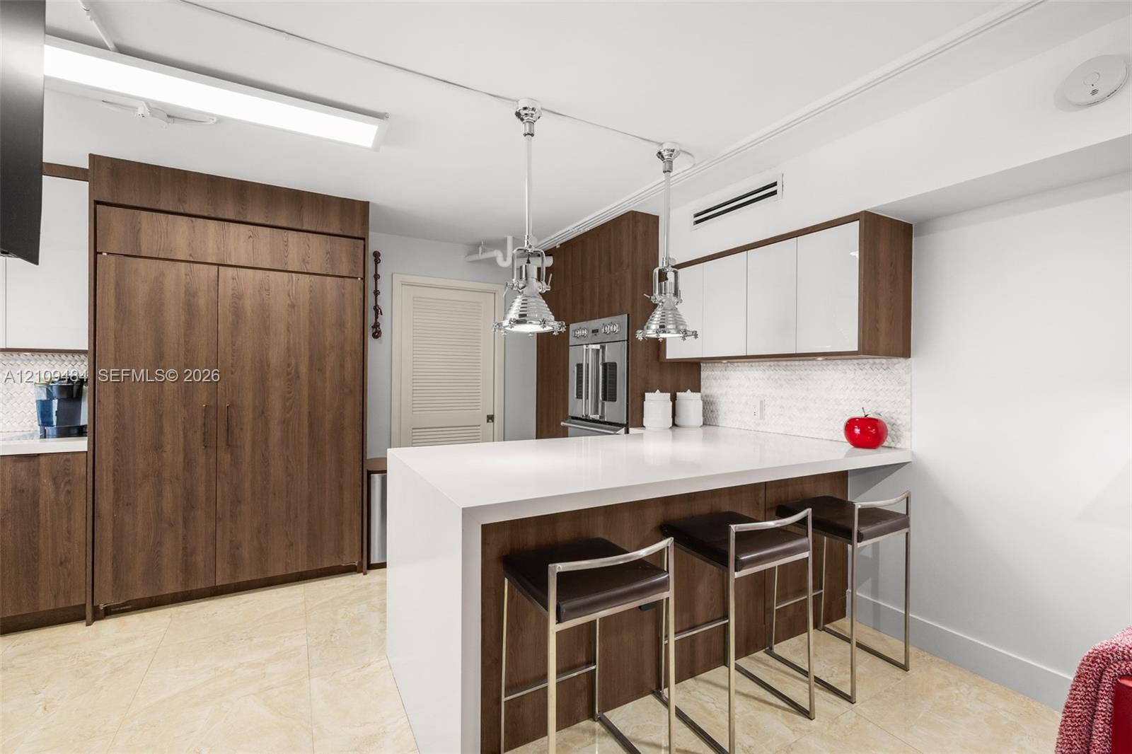 Kitchen featuring a waterfall-edge stone countertop, custom cabinetry with wood-finish and white high-gloss doors, stainless steel wall-mounted oven, and dual polished-metal pendant light fixtures