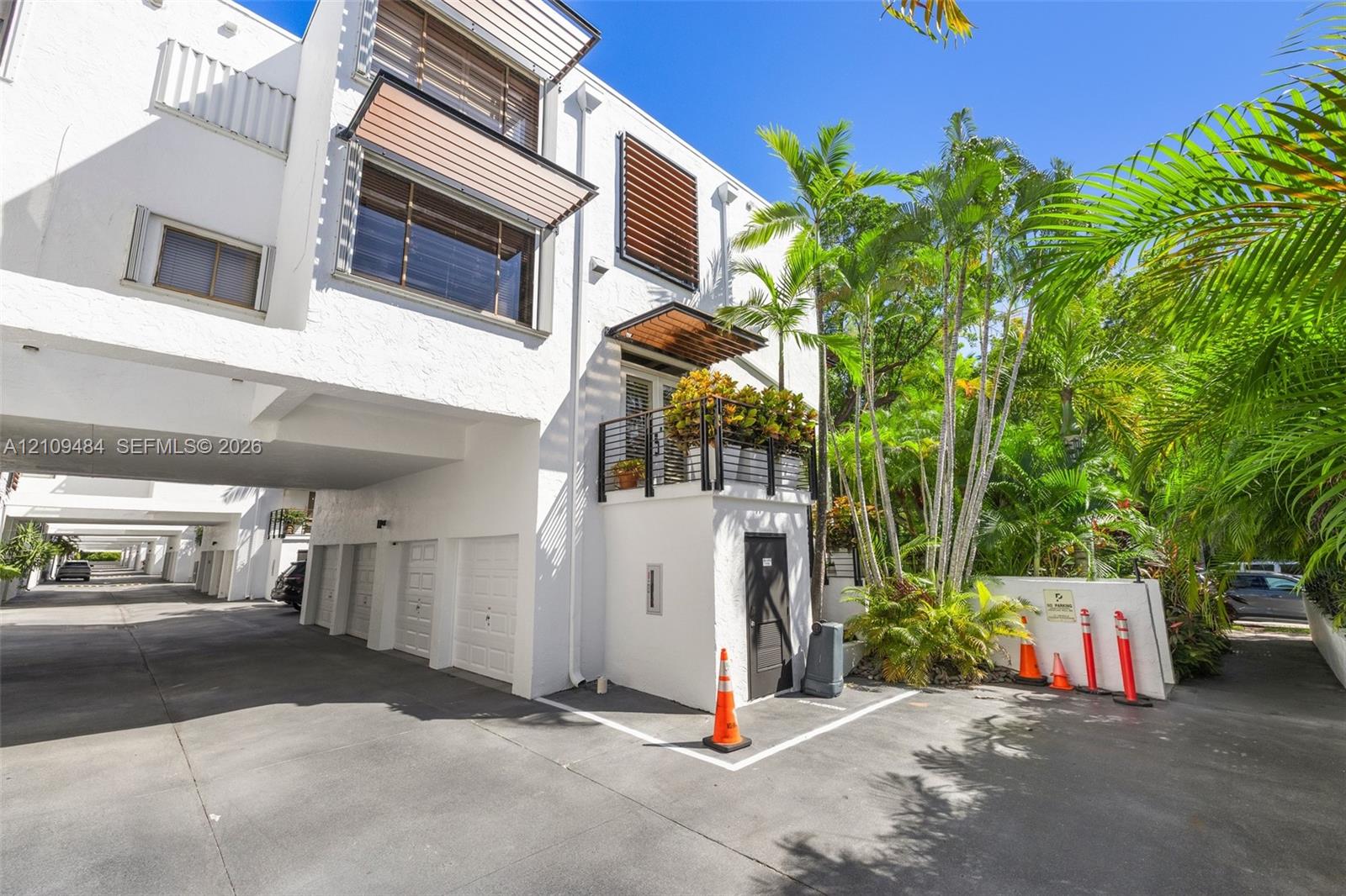 Contemporary multi-level exterior featuring white textured stucco, wood-slat window awnings, elevated balcony with black metal railing, and covered ground-level parking
