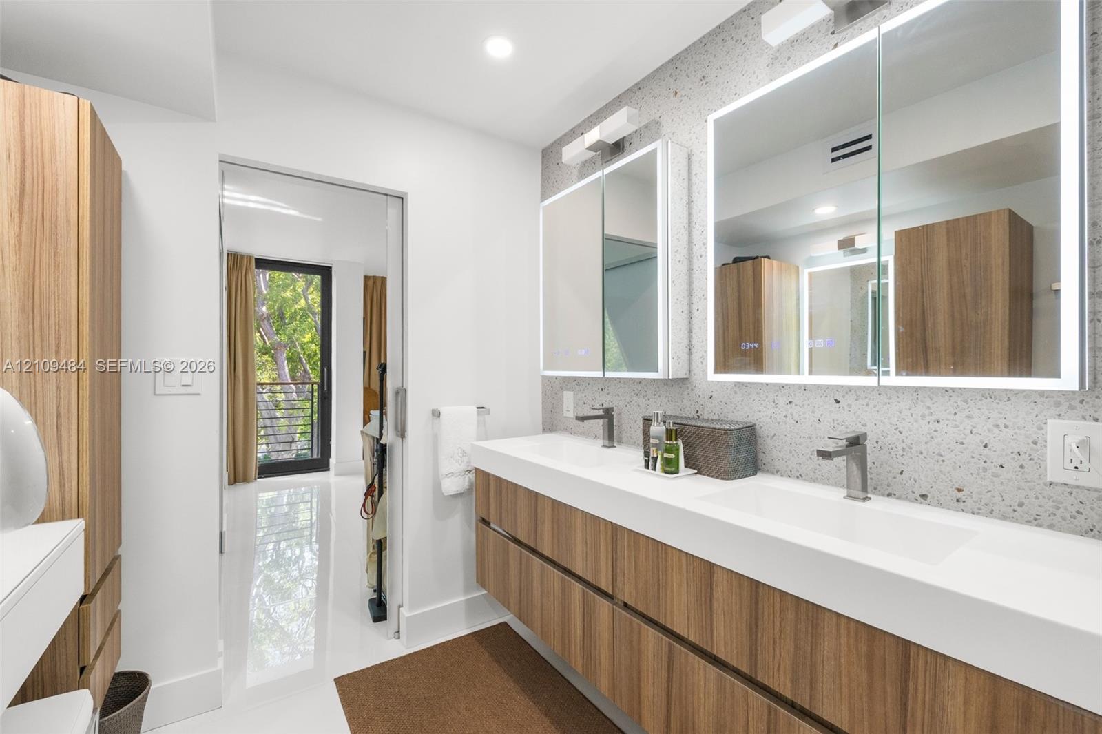 Floating double vanity with wood-finish cabinetry and integrated sinks, speckled stone-finish wall cladding, and dual recessed medicine cabinets with integrated LED perimeter lighting