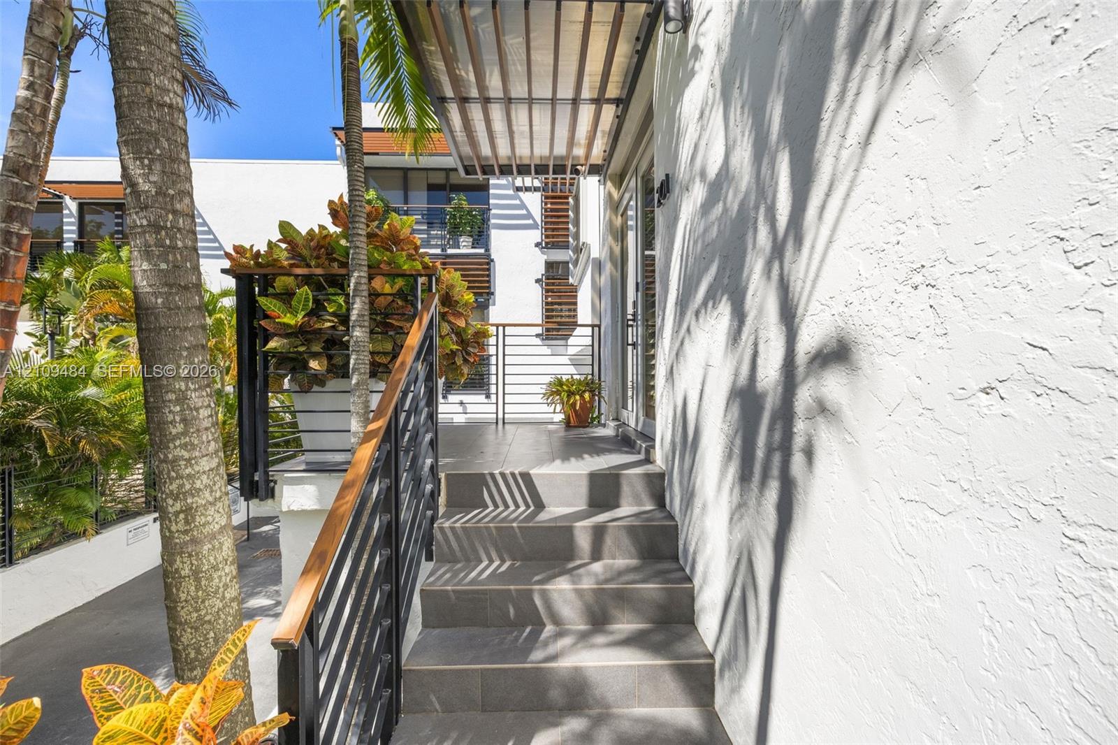 Exterior entryway featuring a modern staircase with tile treads, a wood-accented metal railing, a louvered metal awning, and white textured stucco wall finish