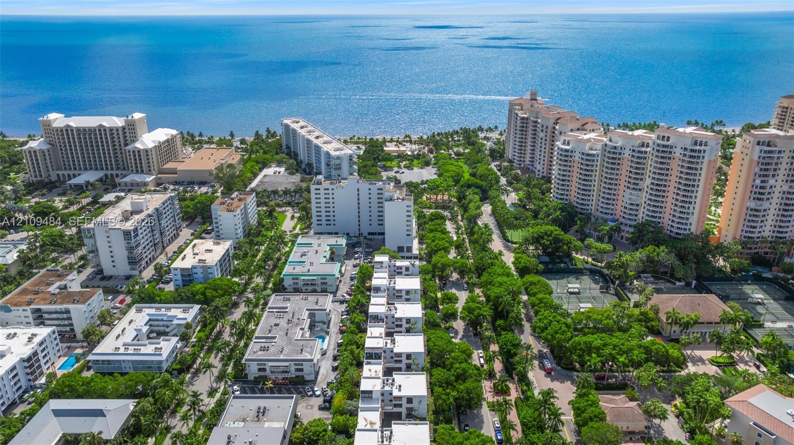 Aerial perspective of a coastal residential community featuring multiple high-rise structures, landscaped green space, tennis courts, and immediate proximity to the ocean