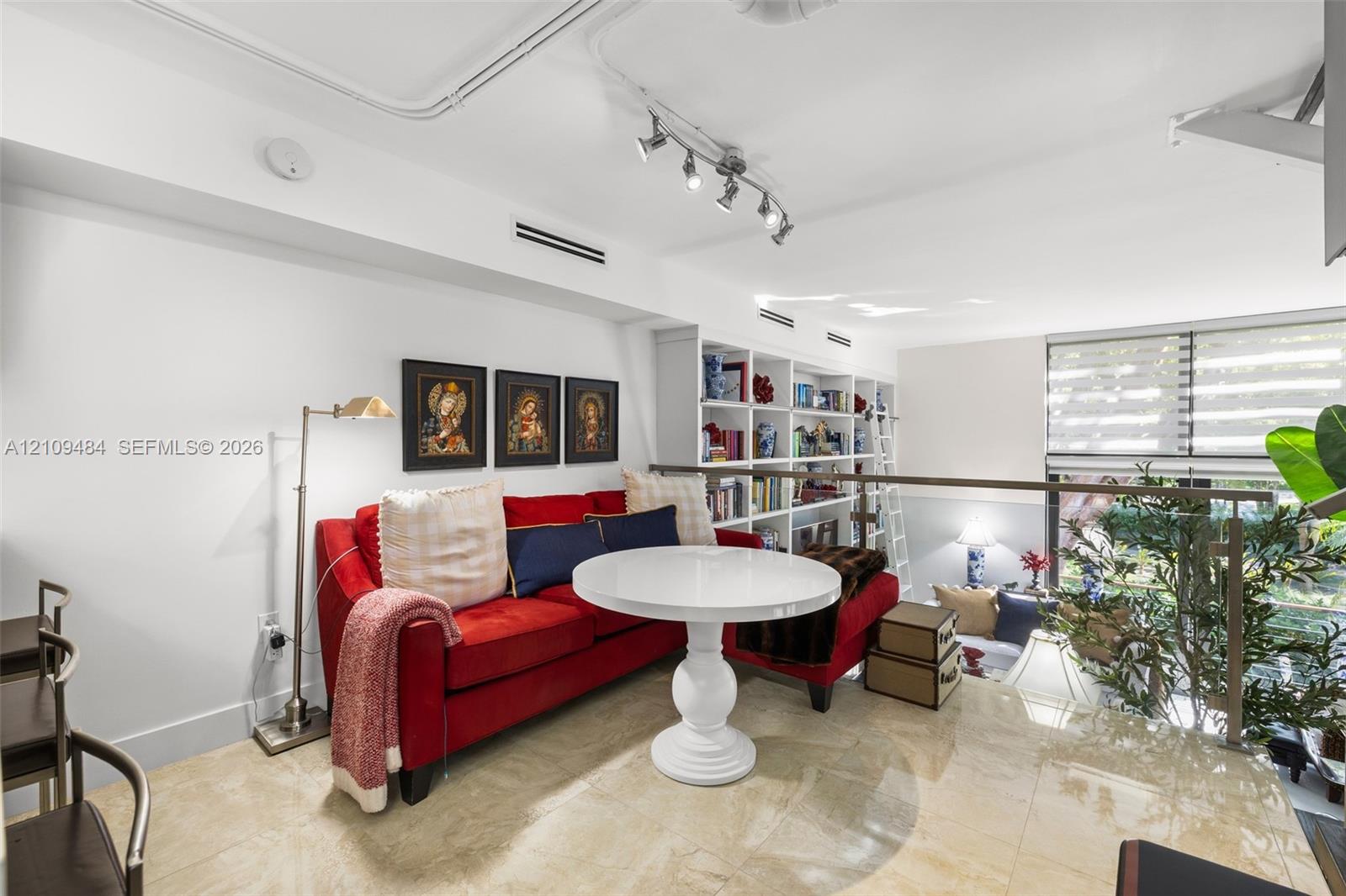 Open-concept interior featuring polished stone flooring, custom built-in floor-to-ceiling shelving with rolling library ladder, adjustable multi-head track lighting, and a metal-framed glass railing