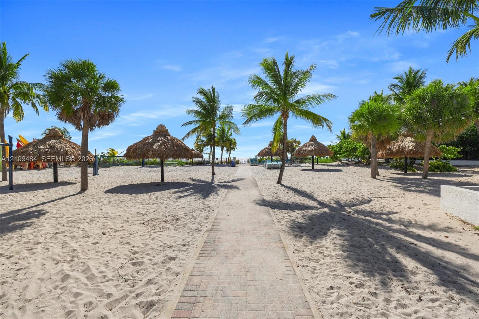 Sandy grounds featuring a central brick paver walkway, multiple thatched-roof palapas, and mature palm trees