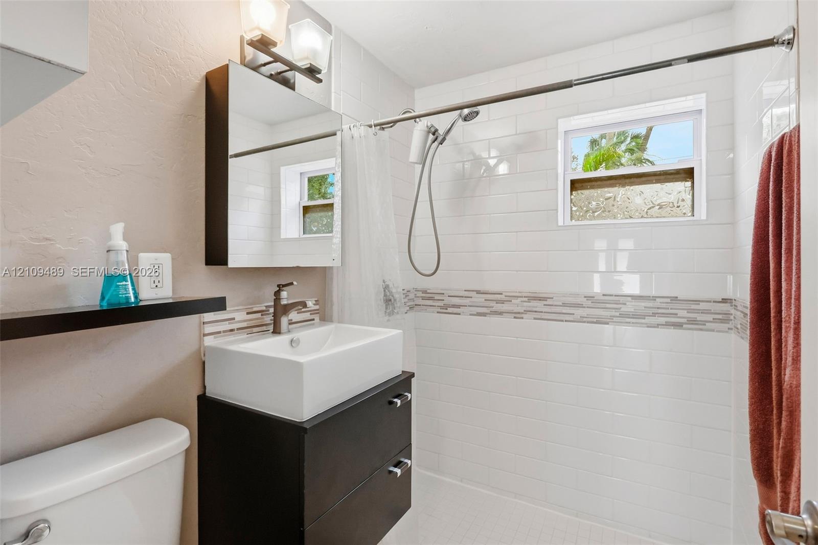 Modern vanity featuring a rectangular vessel sink and matte finish cabinetry, paired with a subway-tiled shower surround accented by a decorative glass mosaic border and a polished metal hand-held shower fixture