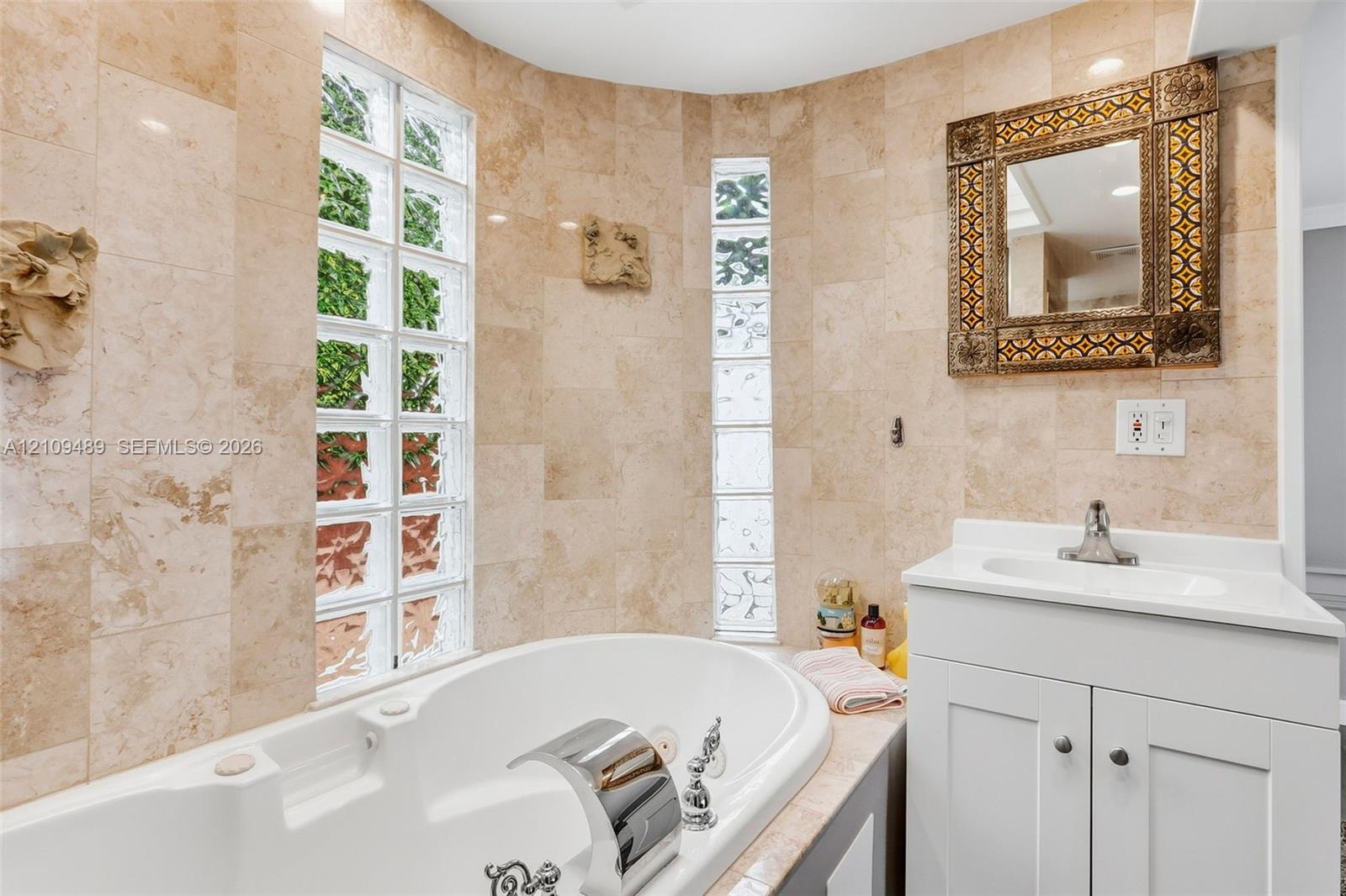 Curved bathtub surround with stone-tiled walls, glass block windows, built-in soaking tub, chrome waterfall faucet, and white vanity with solid-surface countertop