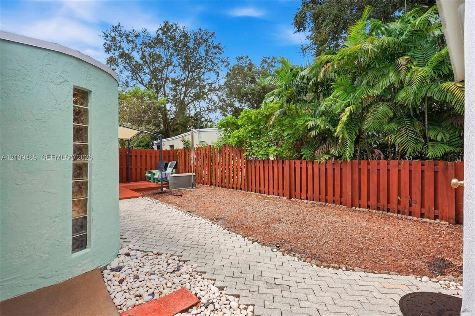 Backyard featuring a curved exterior wall with a vertical glass block window, stone paver pathway, wood-style privacy fencing, and mature palm trees