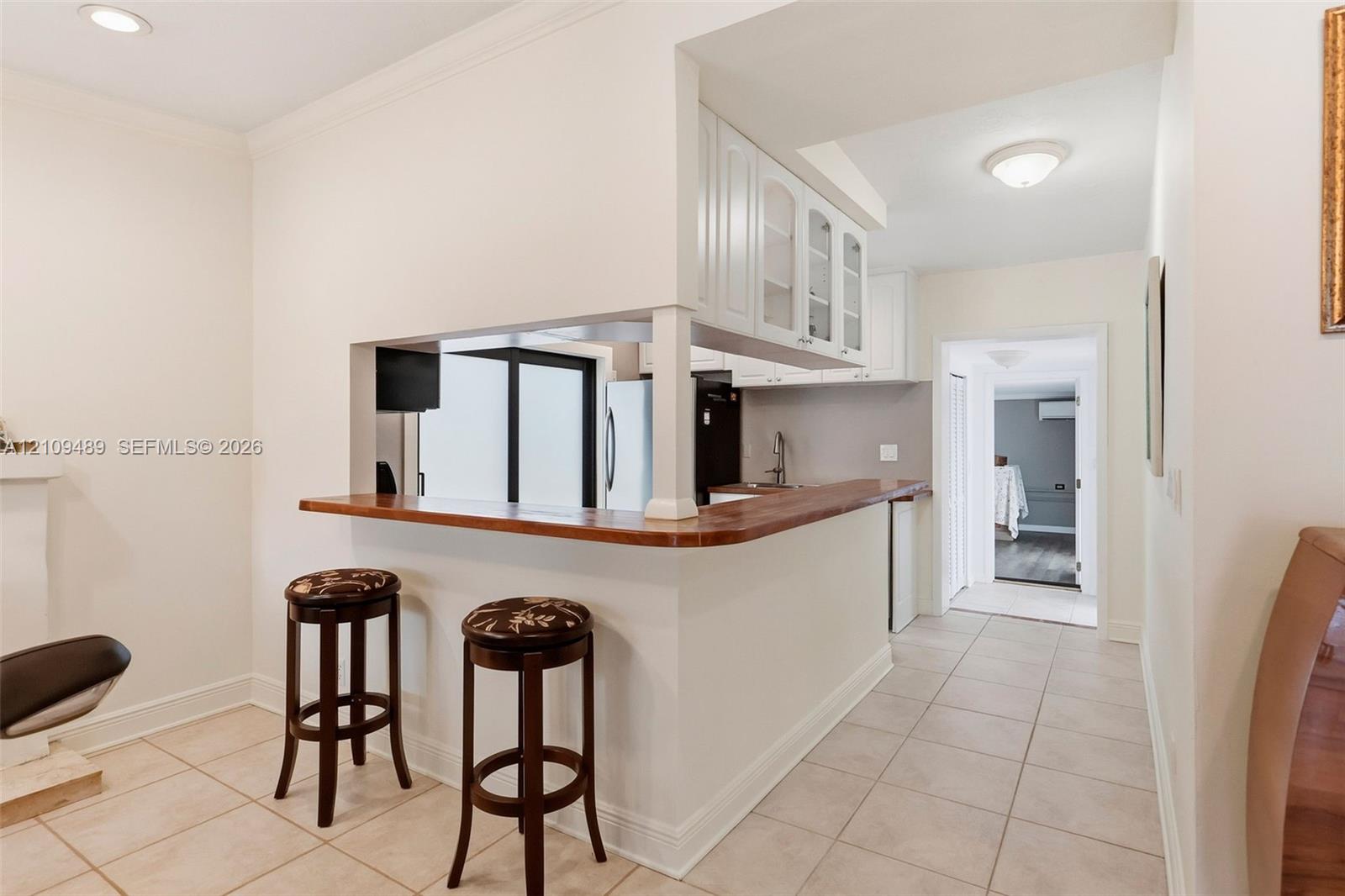 Pass-through kitchen counter with wood surface, white cabinetry with glass-front uppers, and neutral tile flooring