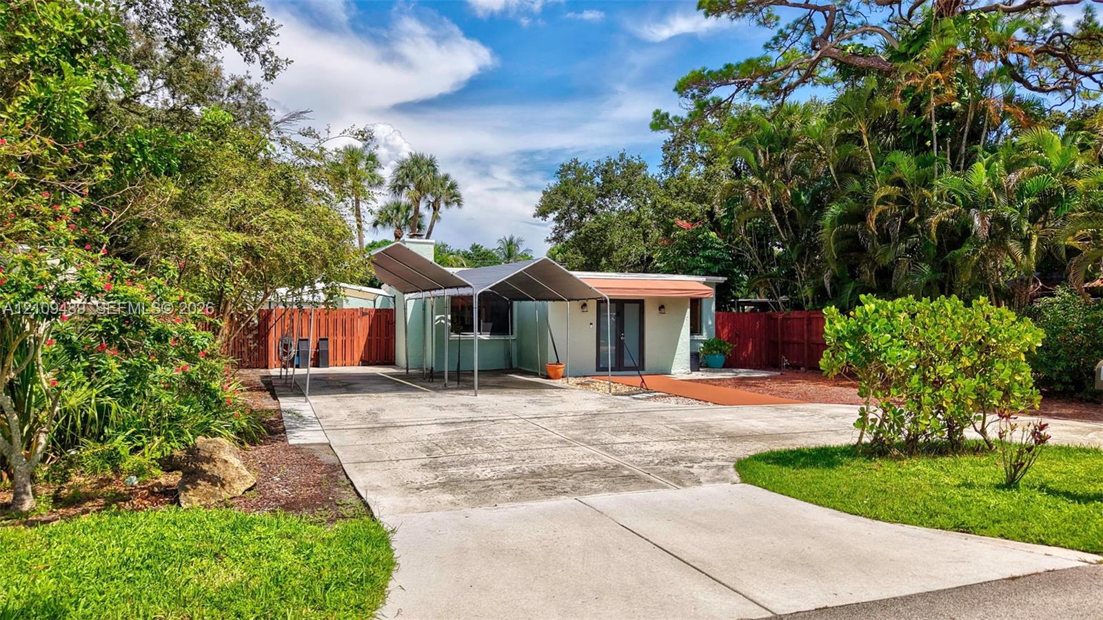 Concrete driveway with a metal carport, horizontal wood-style fencing, and mature landscaping