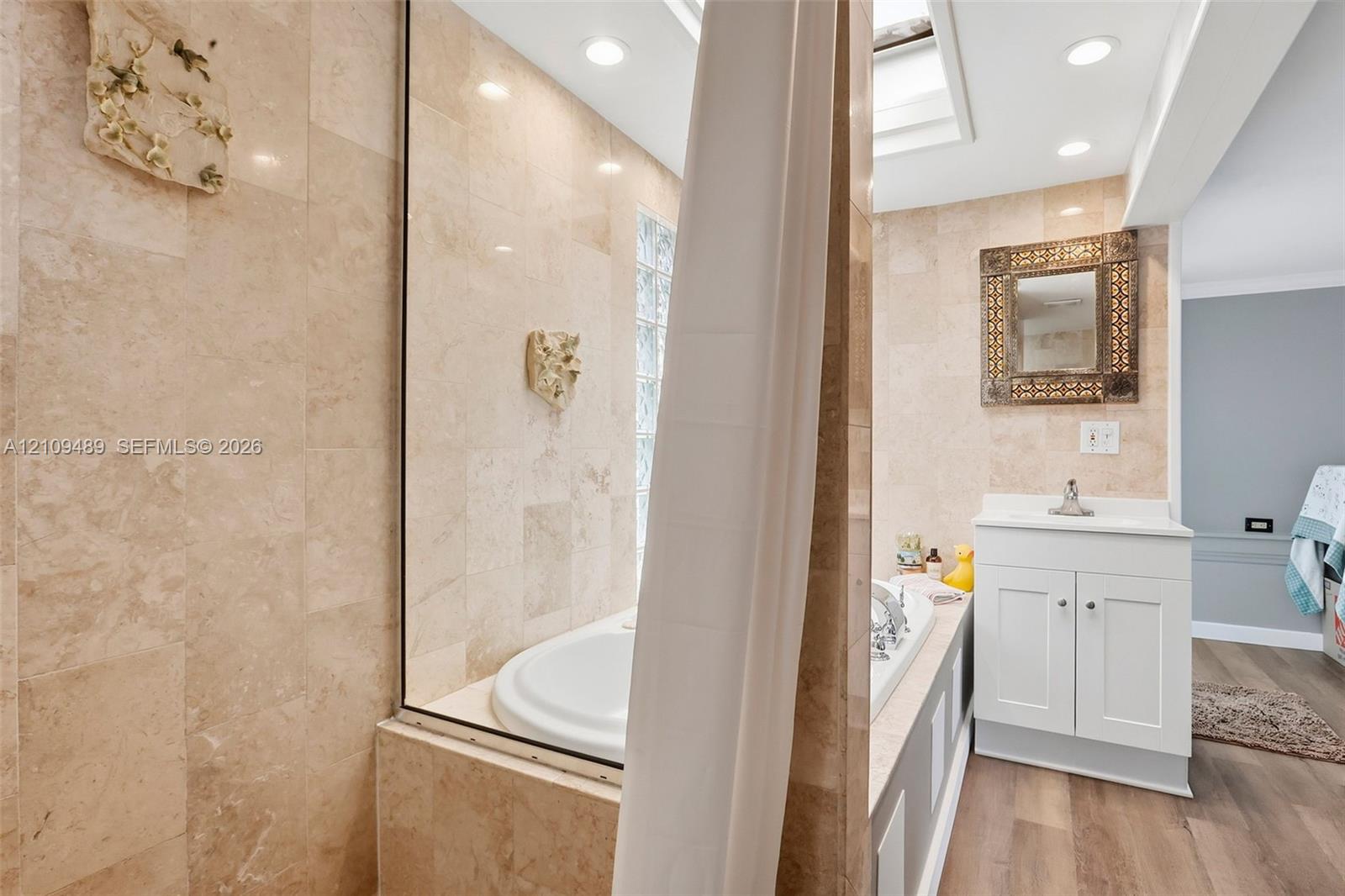 Bathroom featuring stone tile walls, a built-in soaking tub, a skylight, wood-finish flooring, and a white vanity cabinet