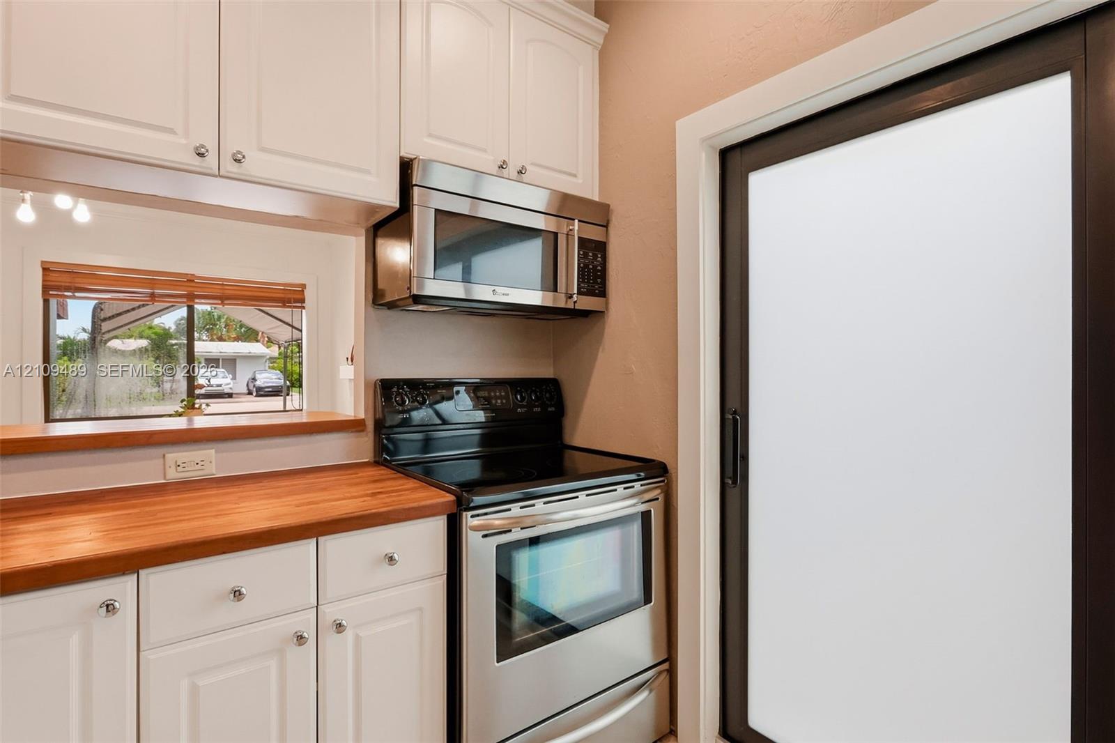 Kitchen featuring white shaker-style cabinetry, stainless steel microwave, black electric range, wood countertop, and a sliding glass door with a dark frame