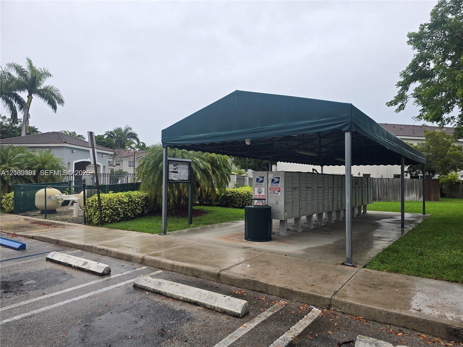 Covered mail kiosk station featuring a dark green canopy, multiple locking postal compartments, and concrete hardscaping