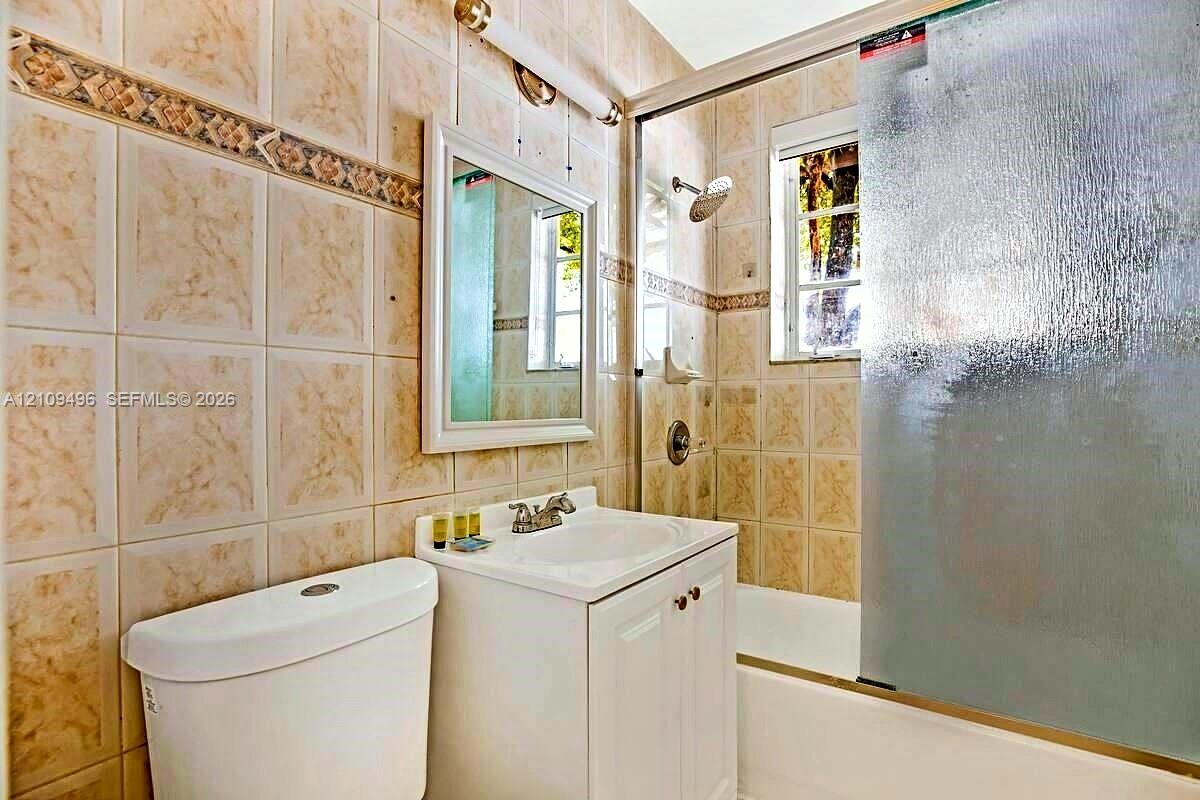 Bathroom featuring floor-to-ceiling tile with decorative accents, white vanity with an integrated sink, polished metal hardware, and a tub-shower combination enclosed by a glass door