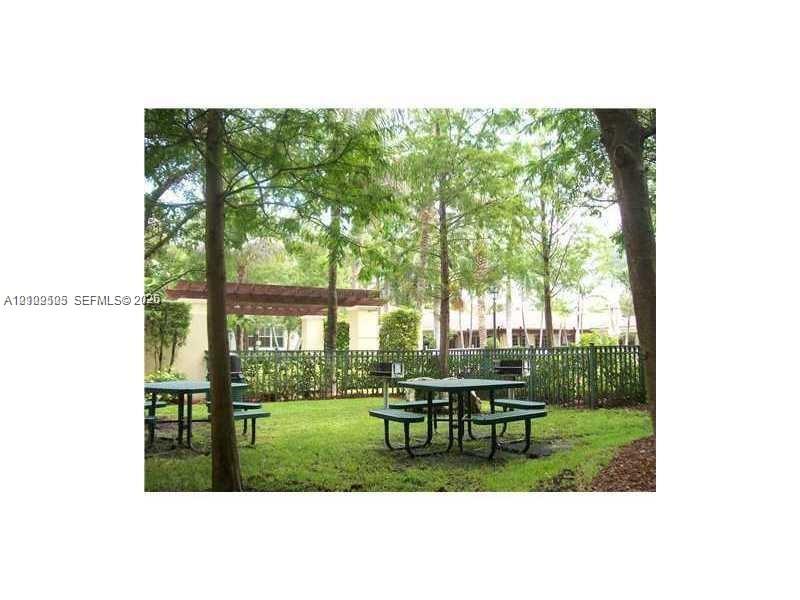 Outdoor recreation area featuring a wooden pergola, built-in metal grills, and mature trees situated on a grass lawn enclosed by a metal fence