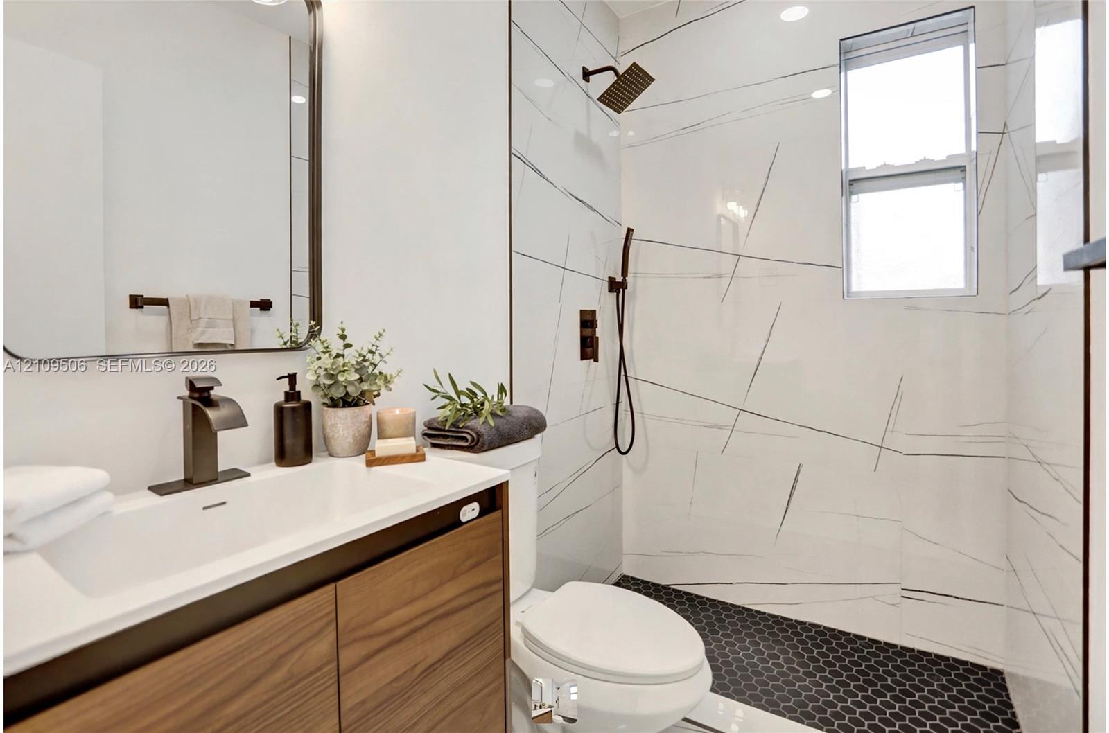 Wood-finish vanity with a rectangular sink, large-format tile shower surround featuring stone-style veining, matte black hardware, and black hexagonal floor tile