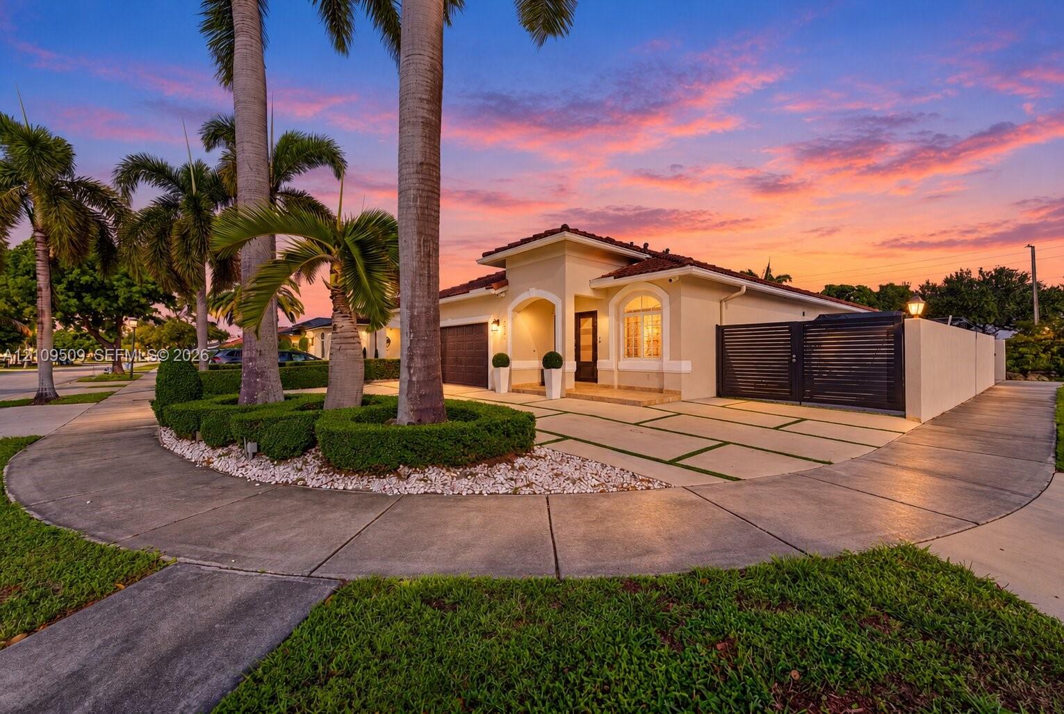 Stucco exterior featuring an arched entryway, segmented concrete driveway with grass inlays, and gated side perimeter wall, complemented by mature palm trees and manicured landscape hedging