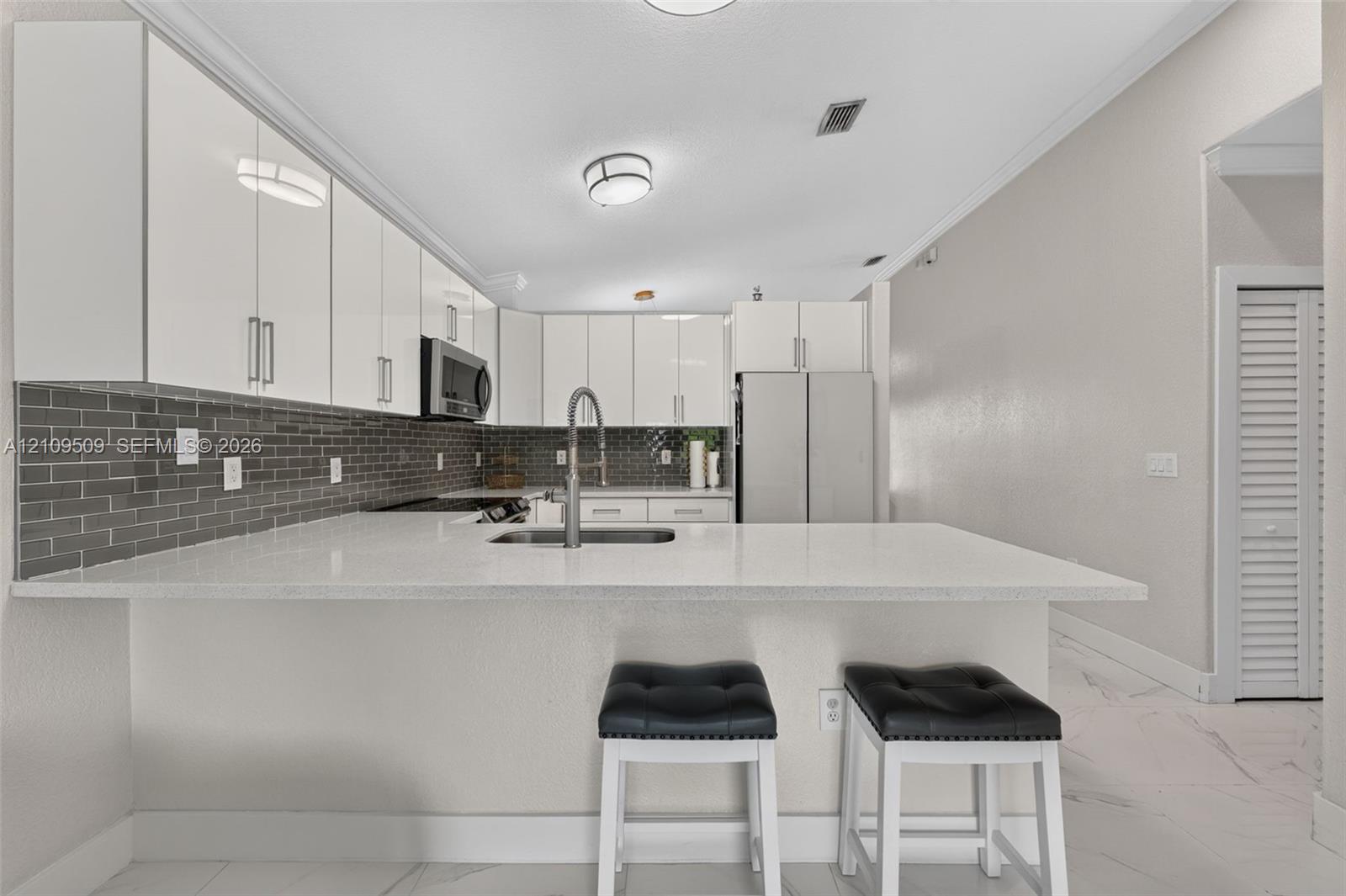 Kitchen featuring high-gloss white cabinetry, white stone-finish countertops, stainless steel appliances, dark grey subway tile backsplash, and light-toned tile flooring