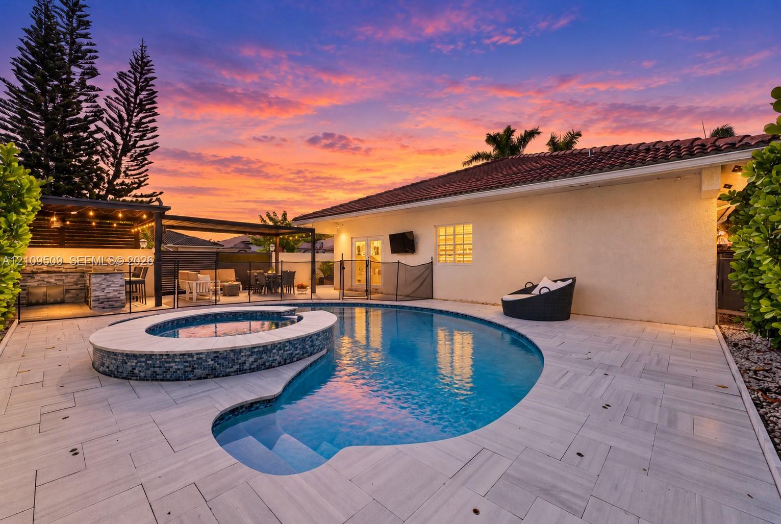 Custom pool with integrated spa featuring mosaic tile detailing, expansive stone-paver patio, and covered outdoor kitchen structure