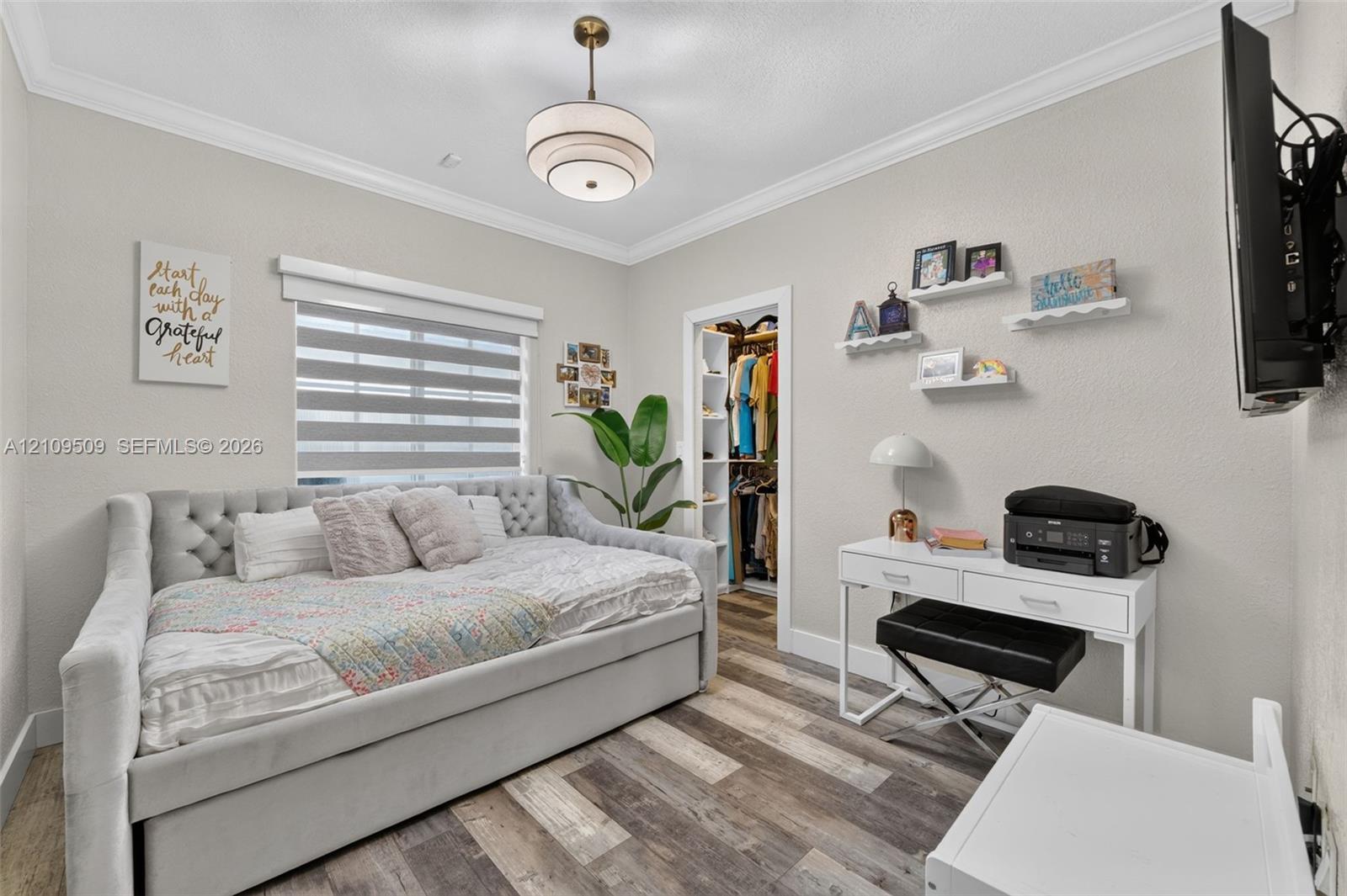 Neutral-toned room featuring wood-finish flooring, crown molding, tiered pendant lighting, a window with roller shades, and a built-in walk-in closet