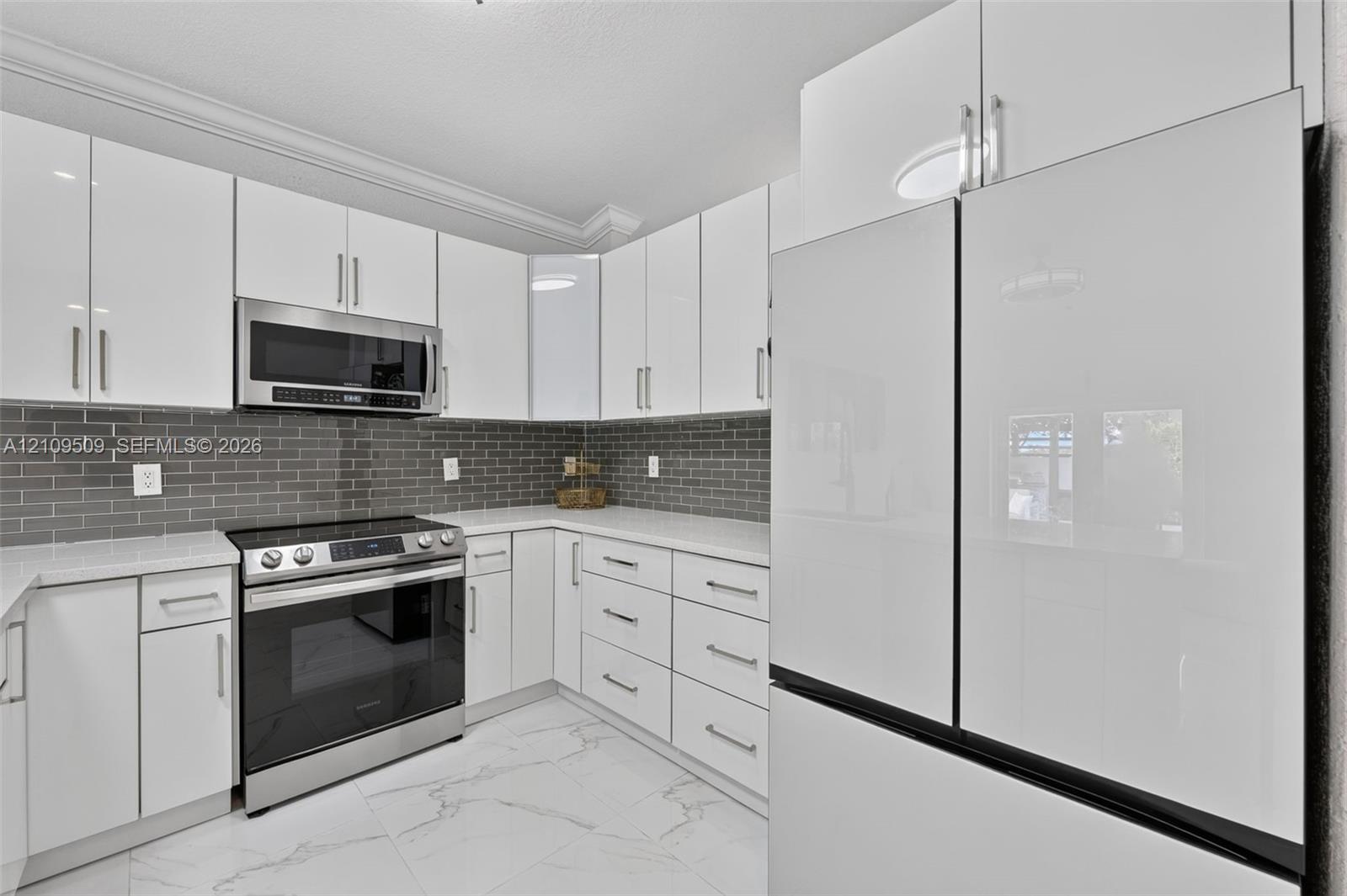 Kitchen featuring high-gloss white cabinetry, light-toned stone-finish countertops, grey subway tile backsplash, stainless steel appliances, and large-format white tile flooring with grey veining