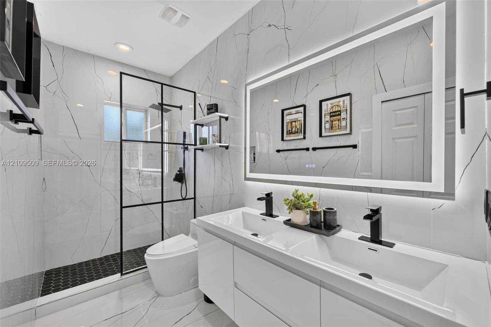 Modern bathroom featuring large-format veined stone tile walls, a dual-sink vanity with matte black faucets, a frameless glass-enclosed shower with black metal grid framing, and a large backlit vanity mirror