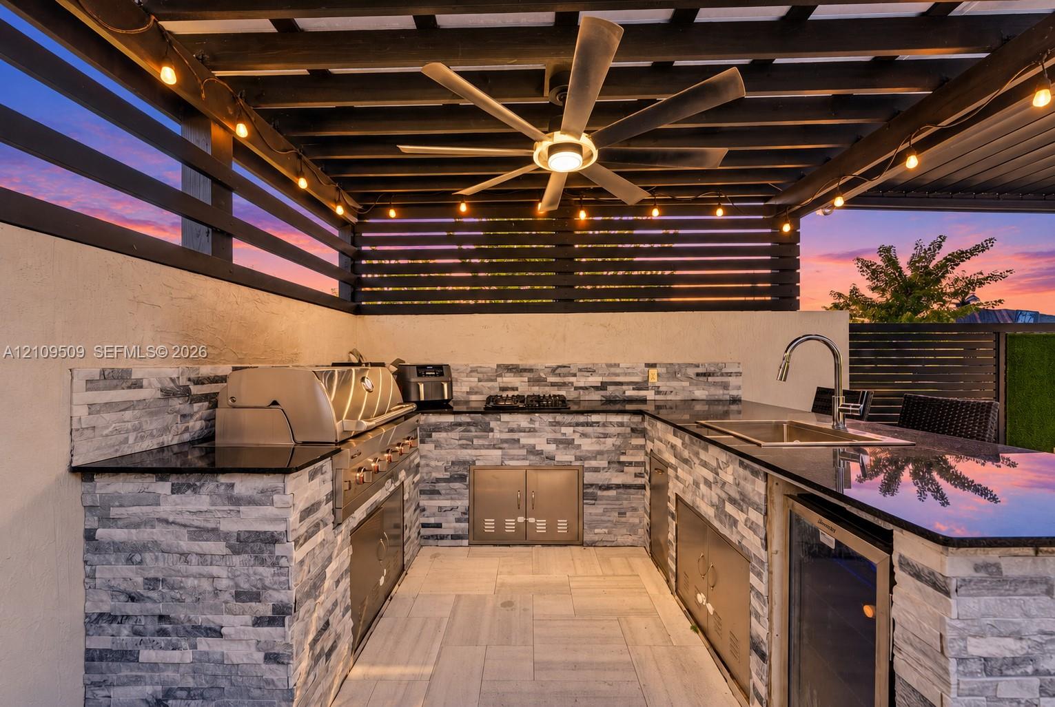 Outdoor kitchen featuring a stone-clad base, dark polished countertops, stainless steel grill, under-counter refrigeration, and a slatted pergola ceiling with a central fan