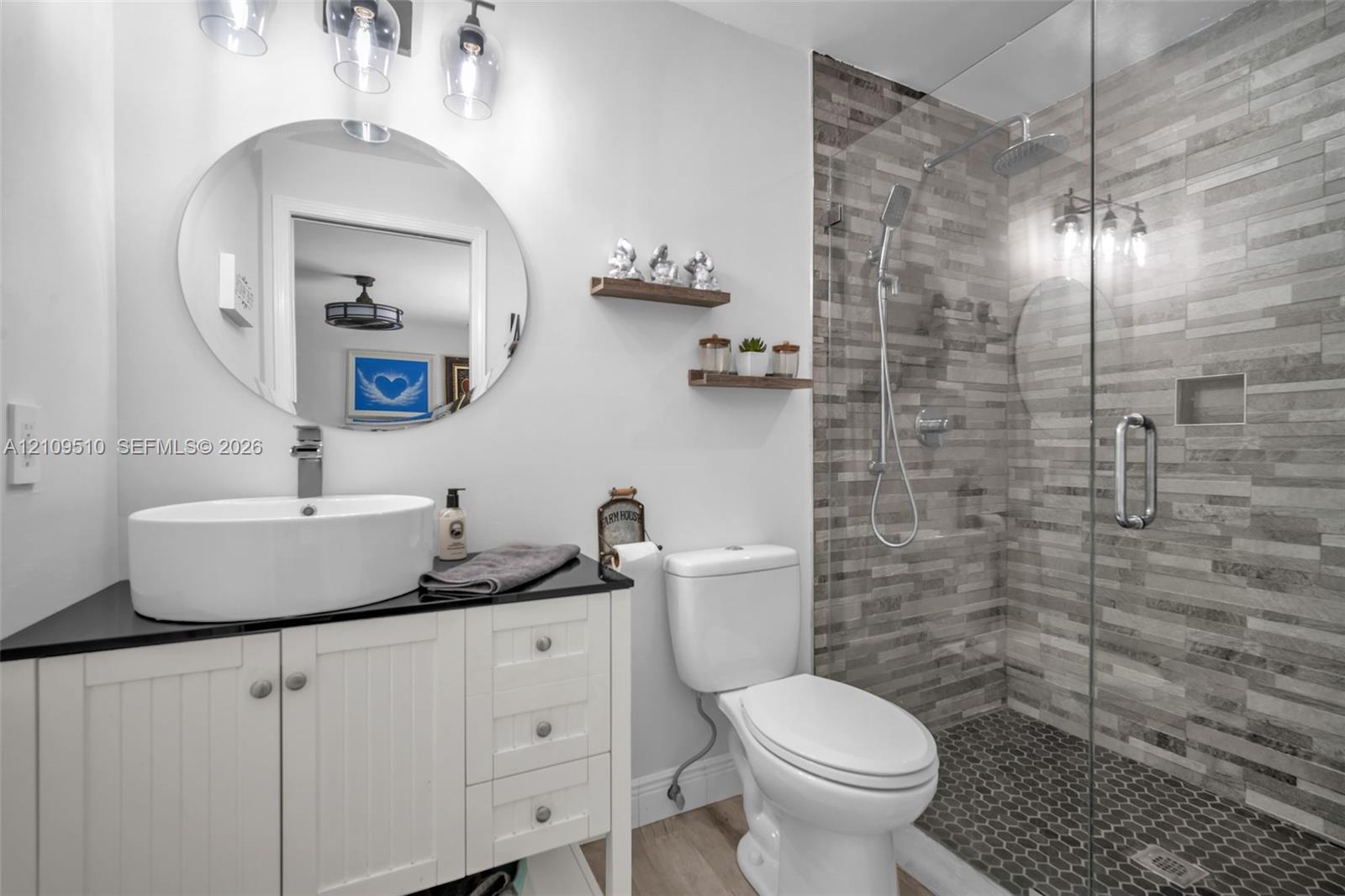 Bathroom featuring a white vanity with a dark countertop, a vessel sink, an oval wall-mounted mirror, and a walk-in shower with textured tile surround and a frameless glass door