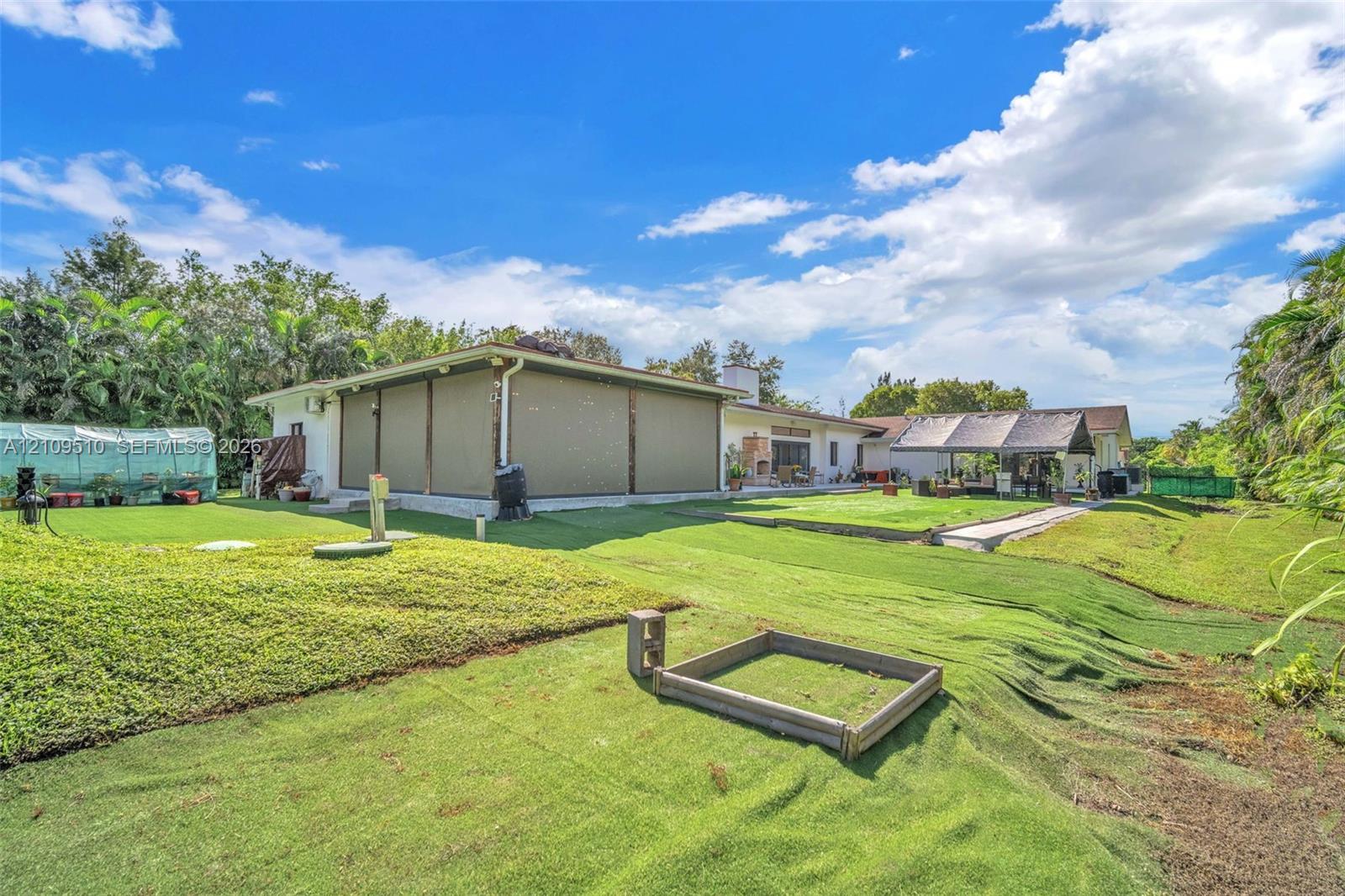 Expansive backyard featuring artificial turf, a permanent greenhouse structure, a covered patio with integrated privacy screens, and a timber-framed garden bed